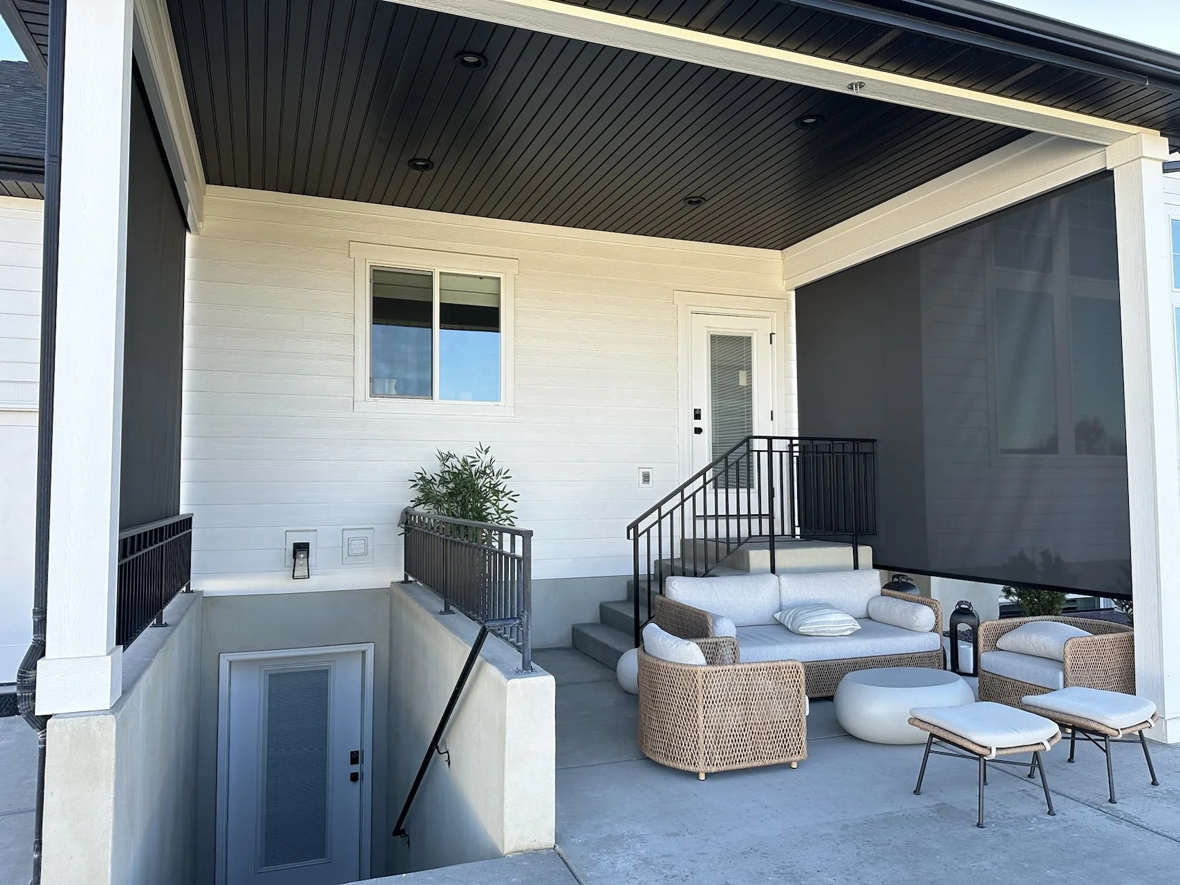 Outdoor patio area with wicker and cushioned furniture, small tables, stairs leading to a door, and a screened area, with a window on the wall.