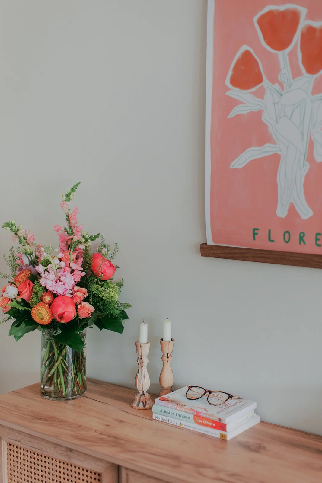 A wooden table with a large glass vase containing pink and natural flowers, two white candles in wooden candle holders, a pair of glasses, and a stack of three books, against a plain light-colored wall with a framed poster of a flower and the word 'FLOUR'.