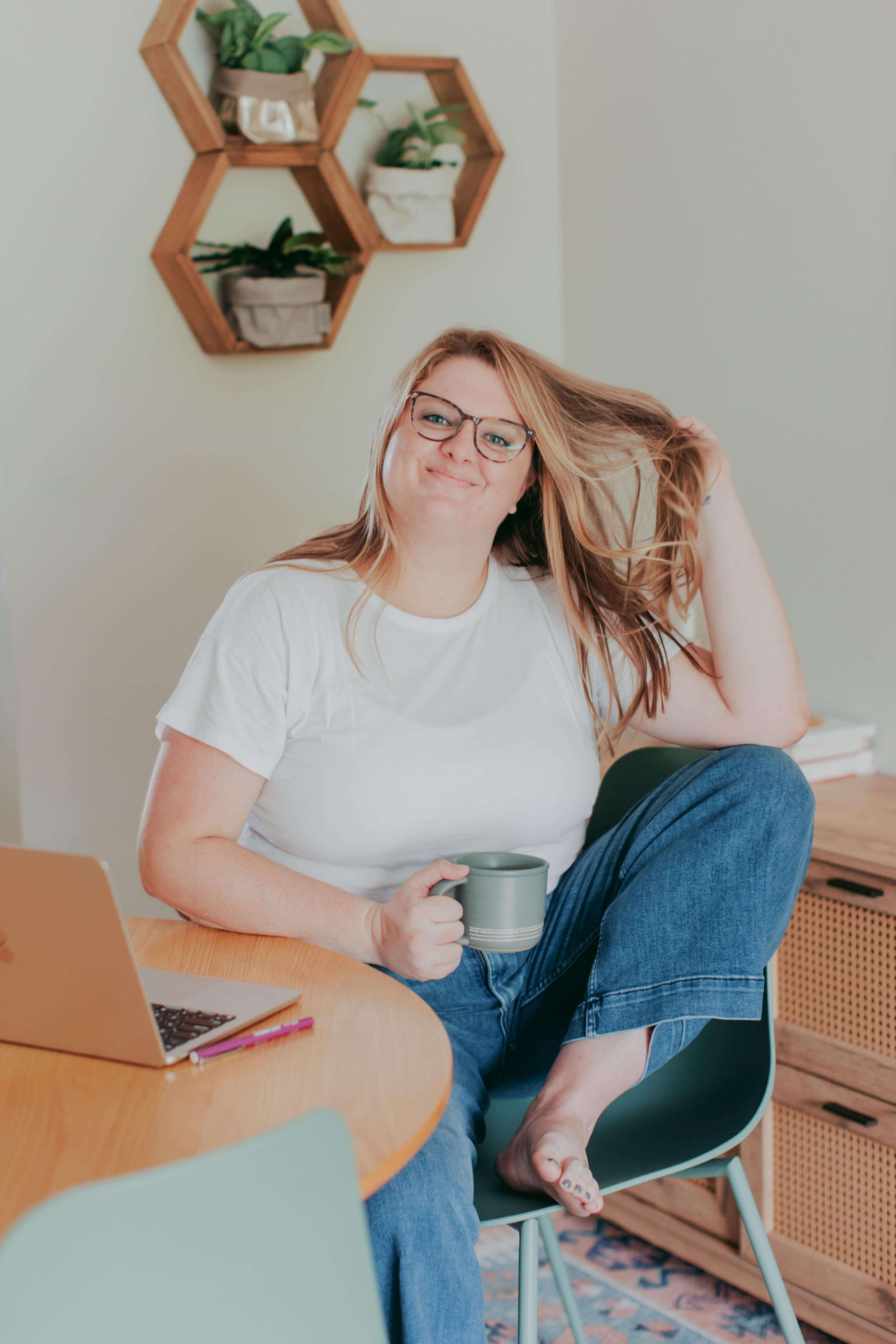 A woman with red hair and glasses sitting at a table, holding a gray coffee mug, with a laptop, pens, and a notebook nearby, in a cozy room with decorative wall shelves.