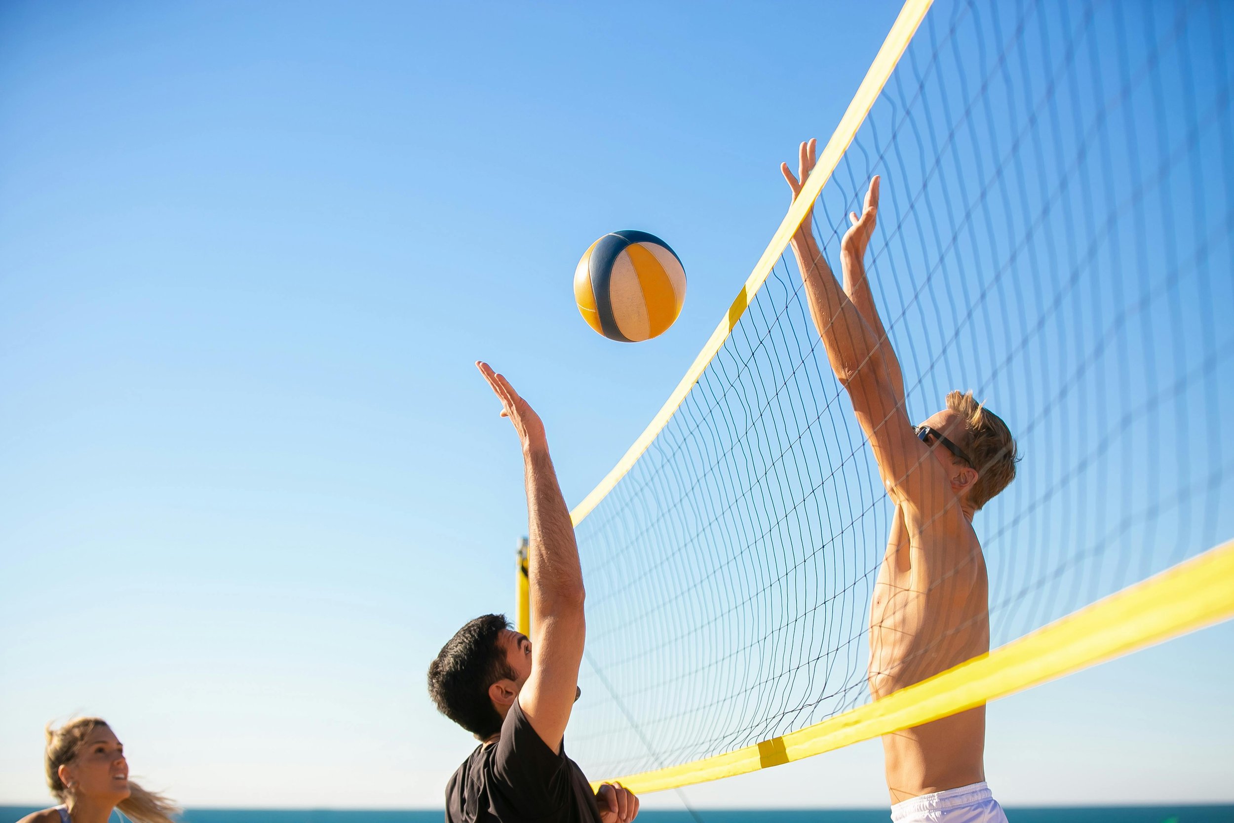 Two men playing beach volleyball at the beach, one spiking the ball while the other jumps to block, with a woman in the background watching.