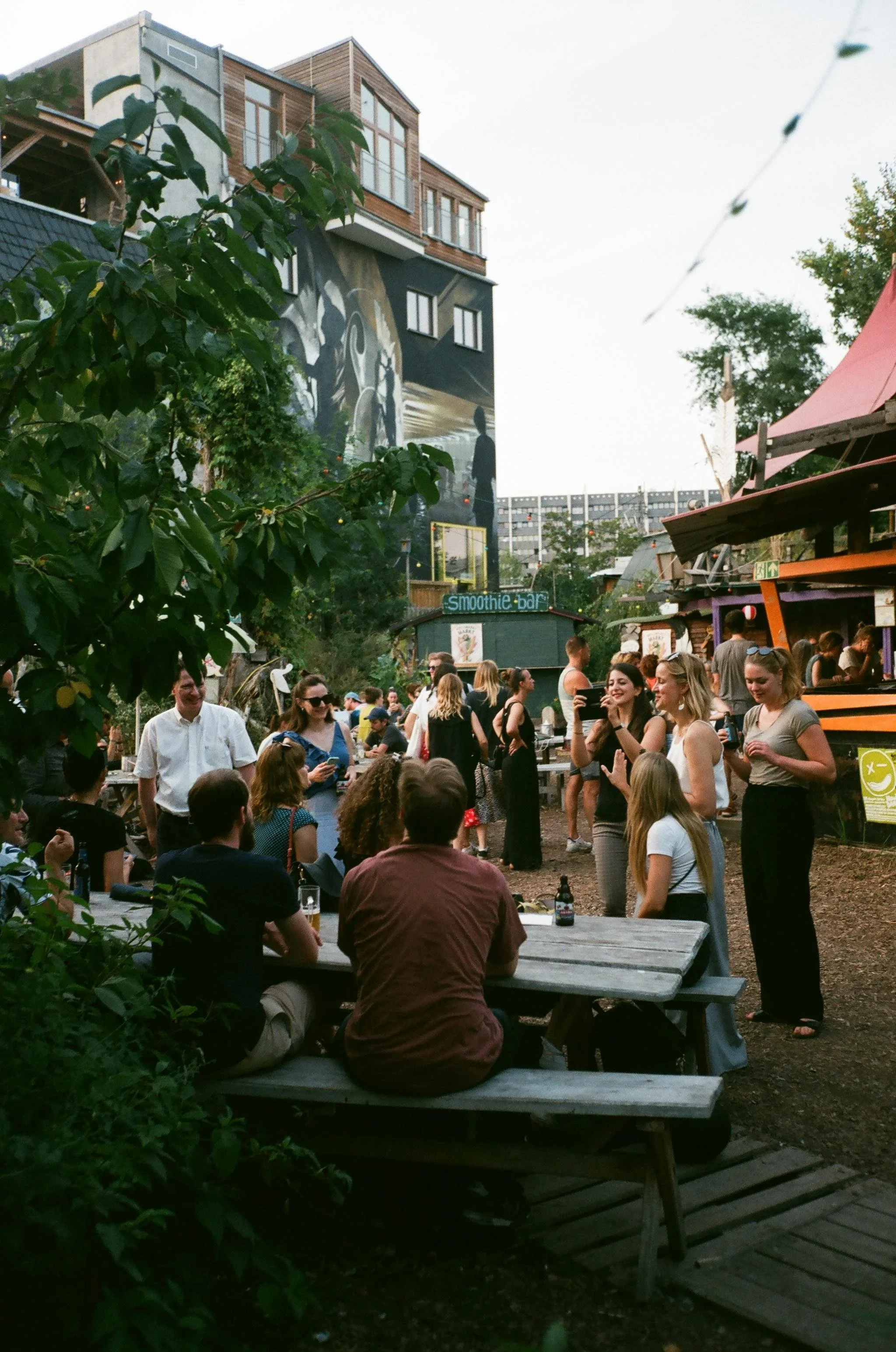 People socializing outdoors at a lively gathering in a backyard or garden area with trees, a mural on a building, a smoothie bar sign, and string lights visible.