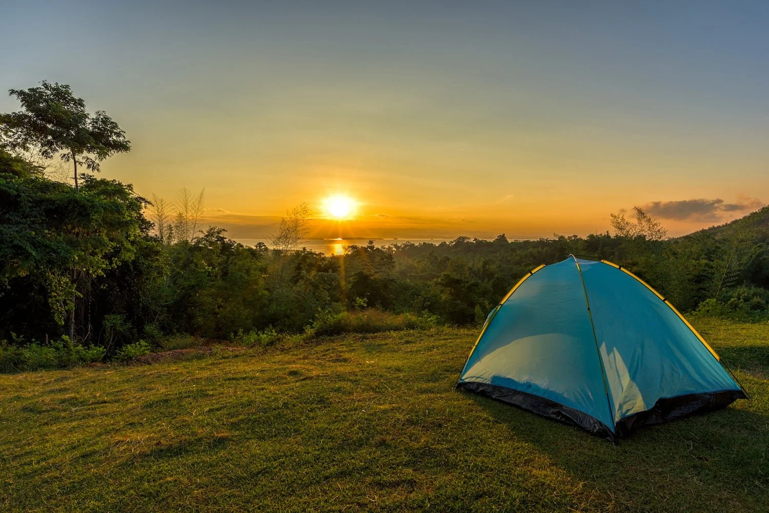 A small blue camping tent set up on a grassy area during sunset, with a forest and a body of water in the background.