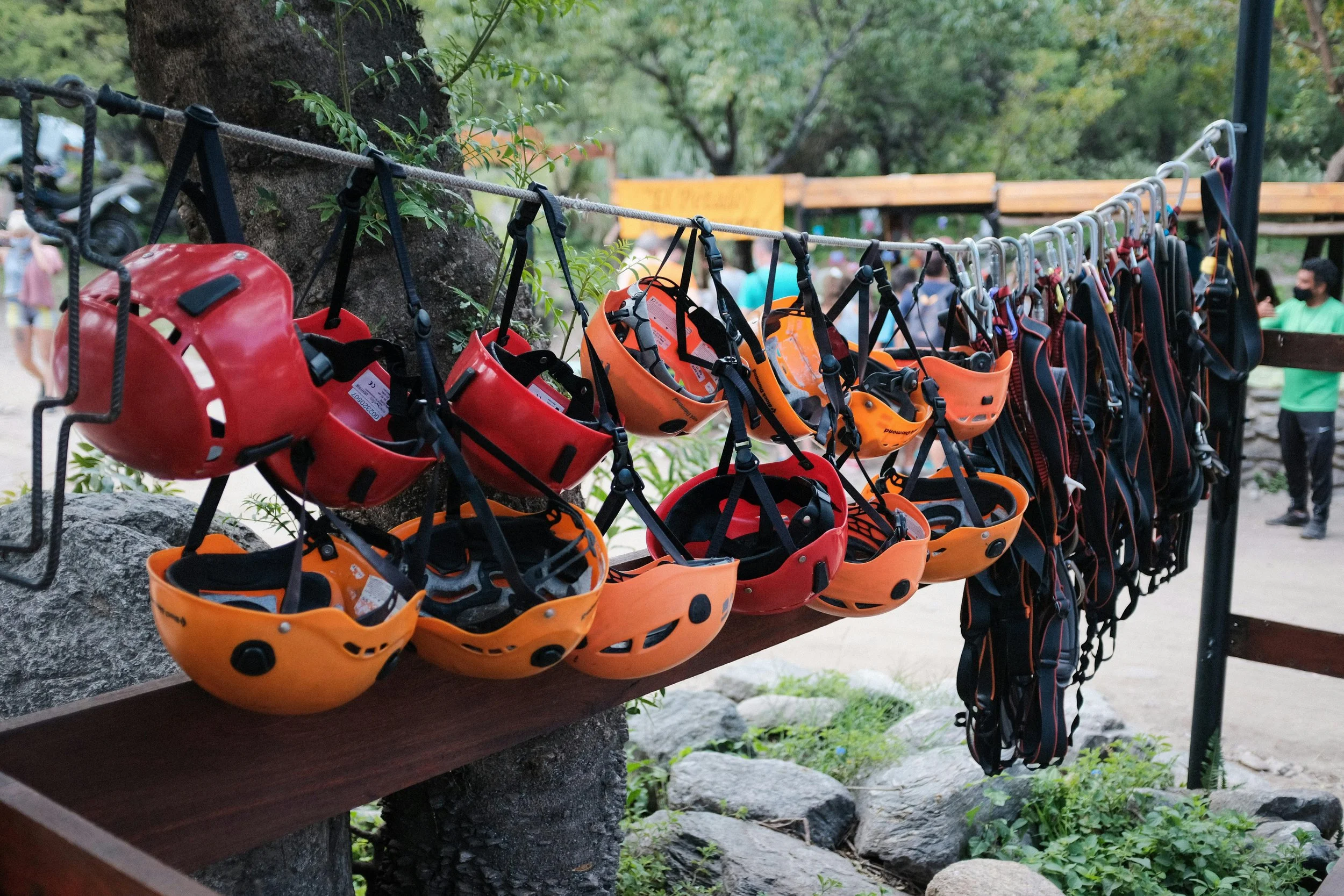 A row of climbing helmets and harnesses hanging on a line at an outdoor climbing area, with trees and people in the background.