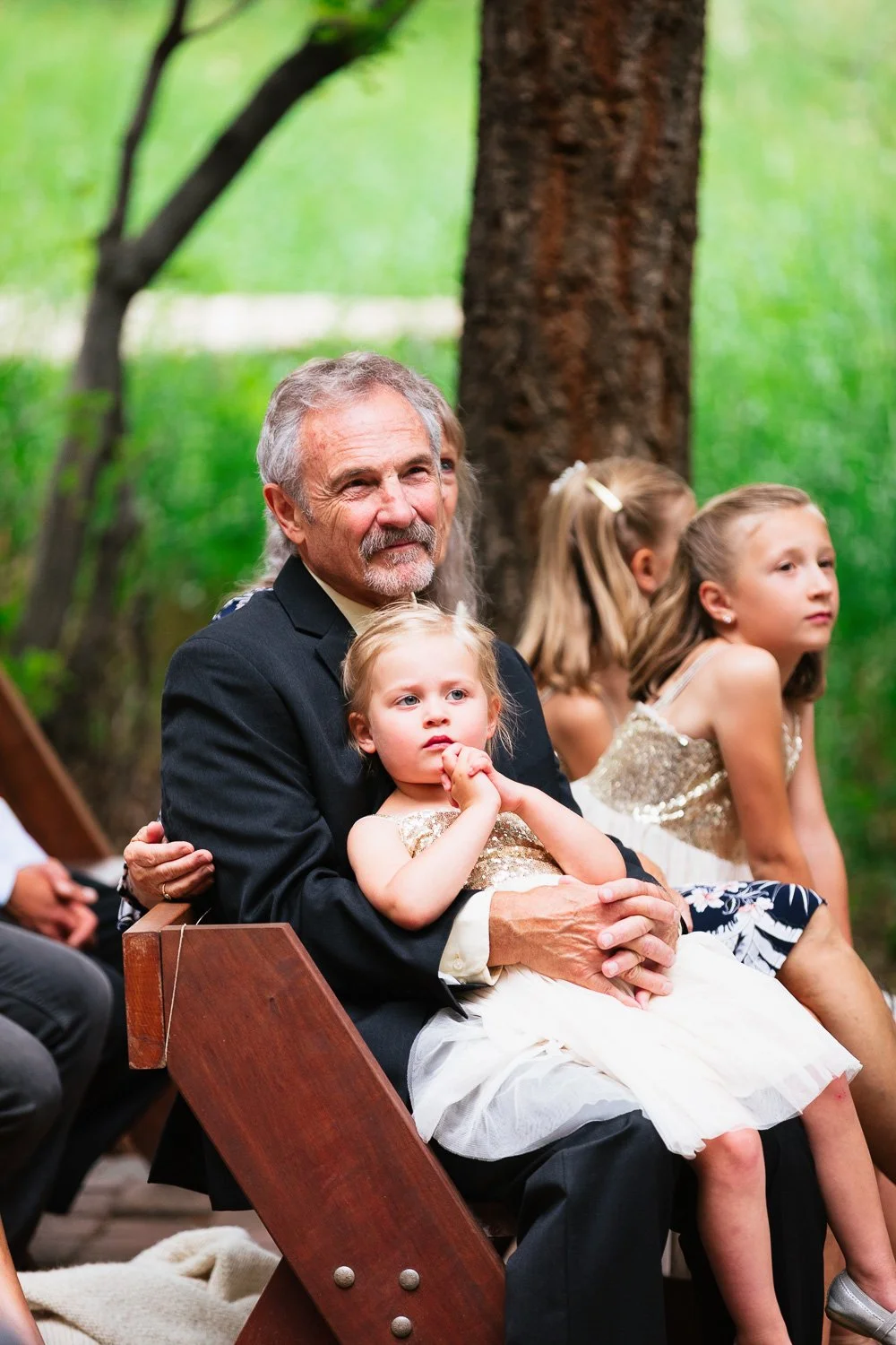An elderly man with gray hair and a beard, dressed in a black suit, sitting on a wooden bench outdoors with young girls. One girl in his lap is wearing a white dress with gold top, looking thoughtful, while two other girls beside him wear dresses with sparkles, sitting on the bench in a forest setting.