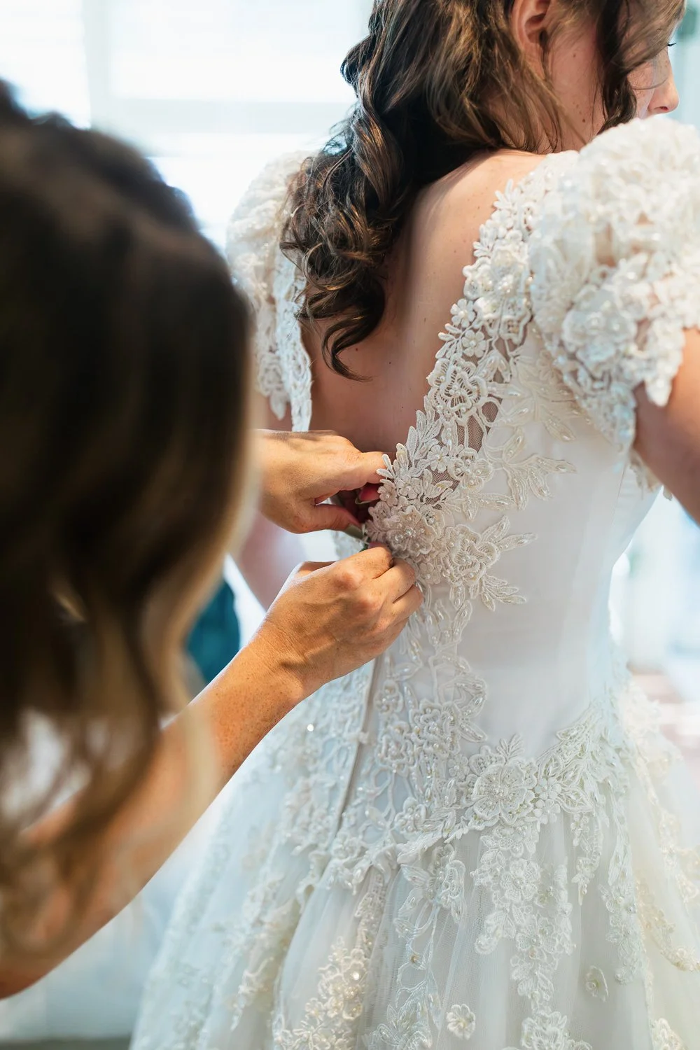 A bride in a white lace wedding dress is being assisted with the back zipper or button by another person.