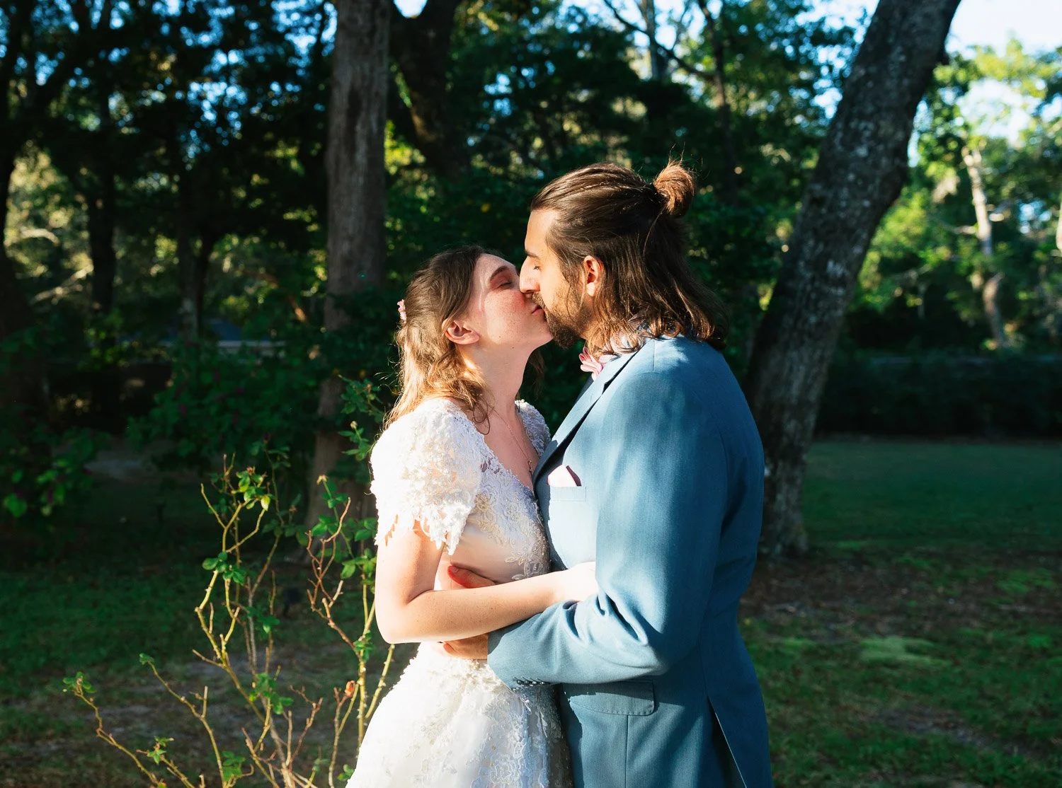 A couple in wedding attire sharing a kiss outdoors in a wooded area.