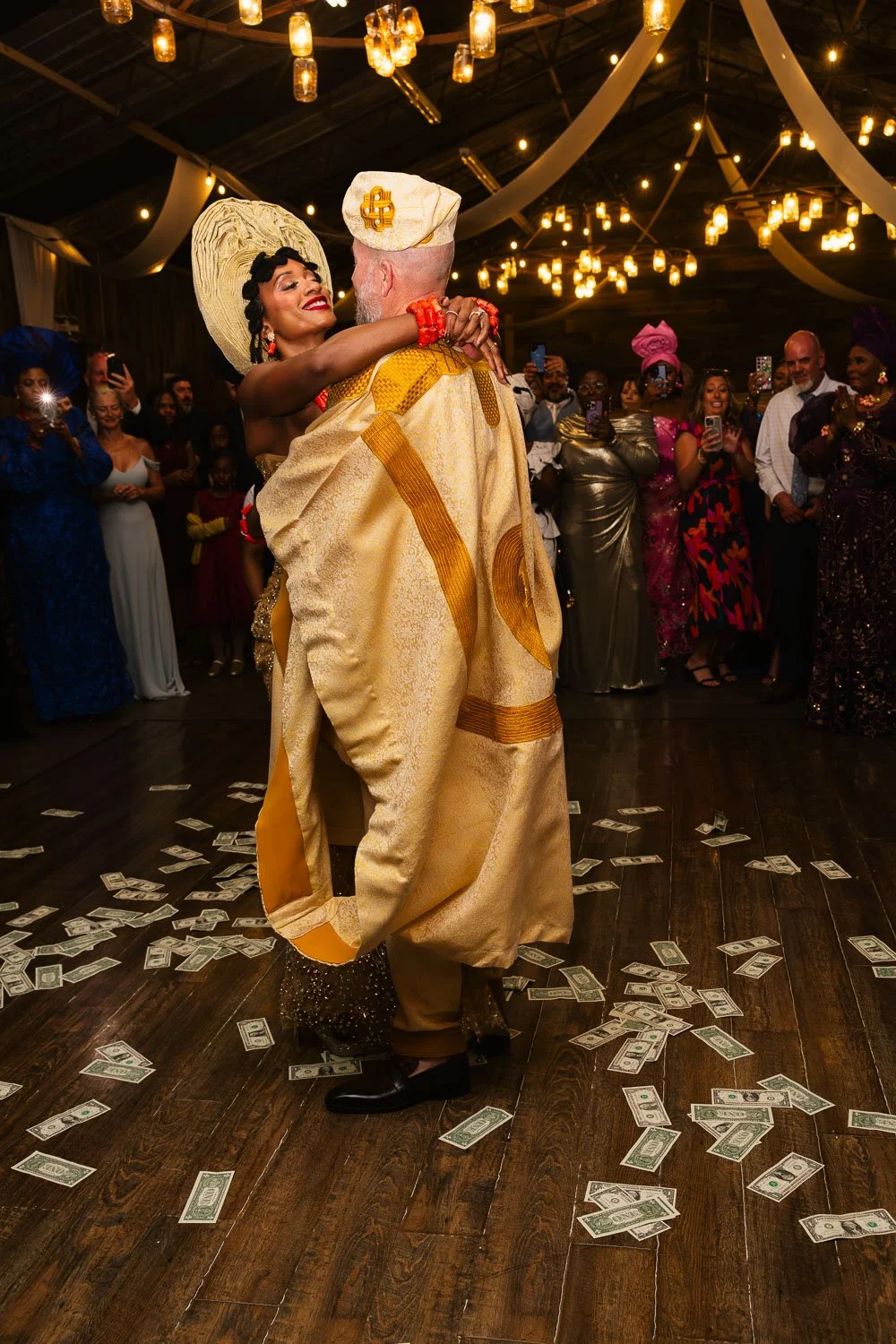 Couple dancing at a celebration with dollar bills scattered on the floor; the man wears traditional African attire and the woman a large hat and glittery dress, surrounded by guests in colorful outfits under warm hanging lights.