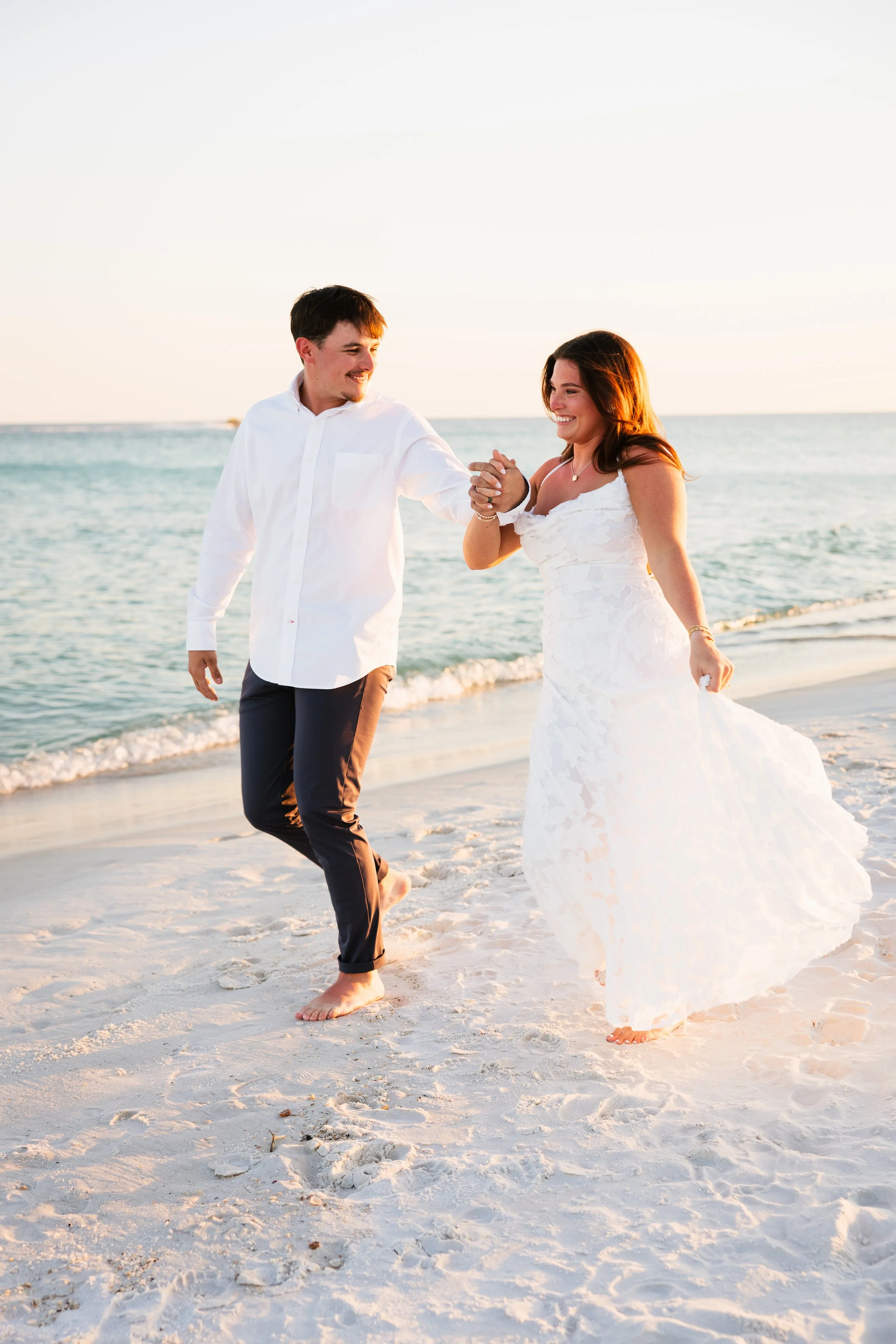 A happy couple dressed in wedding attire walking hand-in-hand on a sandy beach at sunset, smiling at each other with the ocean in the background.