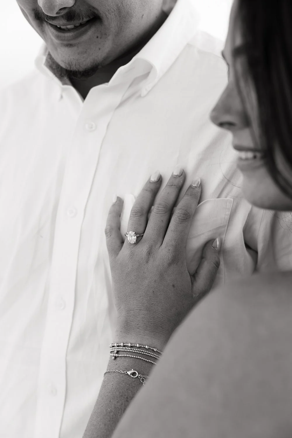 Close-up of a woman's hand with a diamond ring resting on a man's chest. The woman is wearing multiple bracelets. The man is wearing a white dress shirt, and the woman has long dark hair.