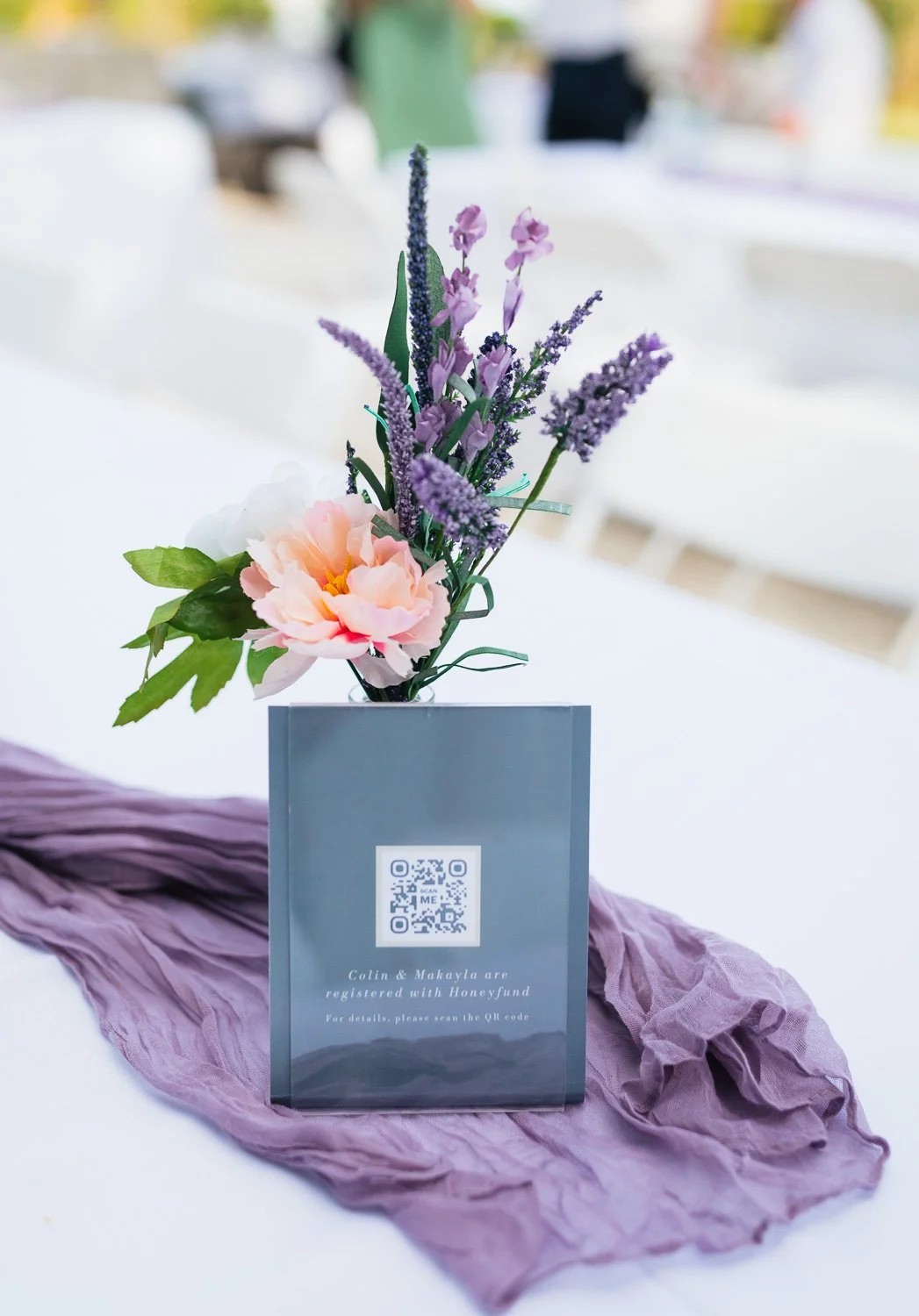 A floral centerpiece with pink and purple flowers in a black vase. A QR code and text are displayed on a gray card in front of the flowers, placed on a purple fabric runner on a white table. The background is blurred.