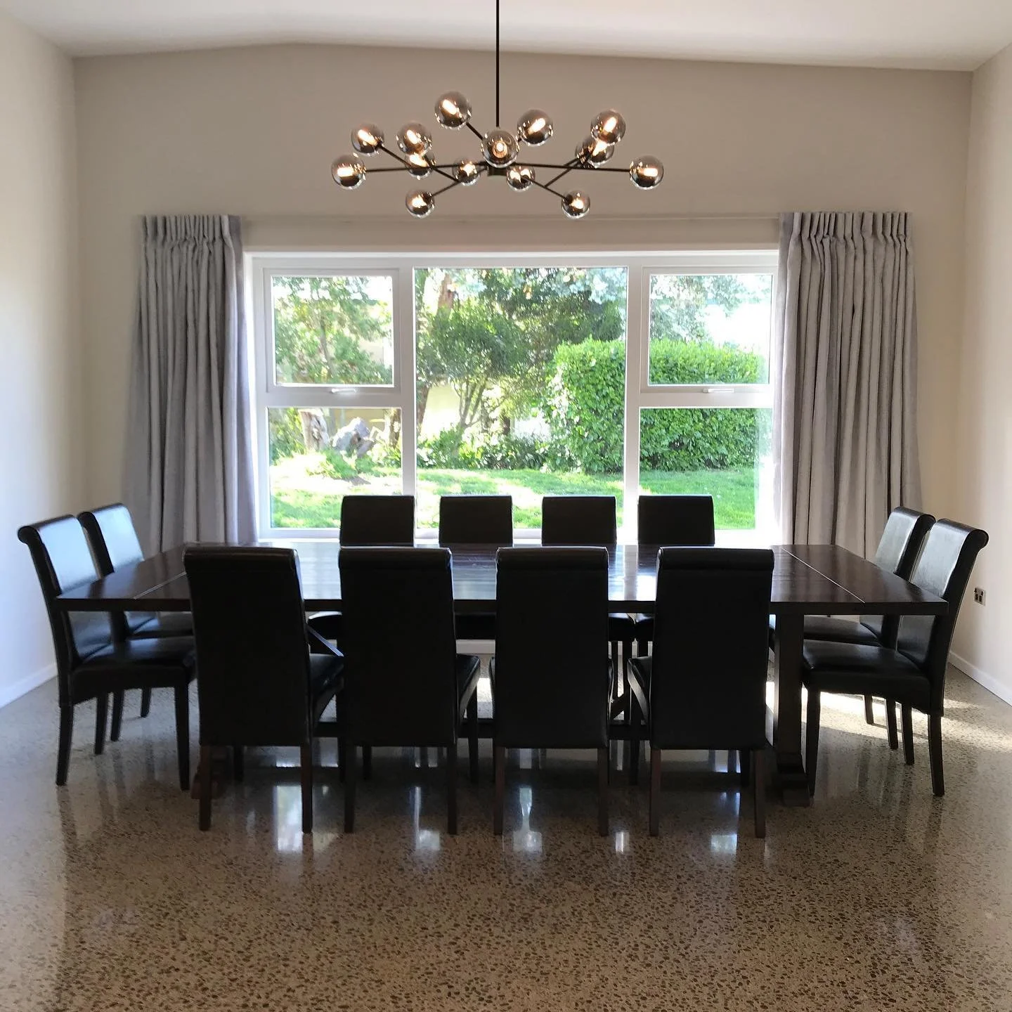 A modern dining room with a large wooden table surrounded by twelve black chairs, a large window with white framing and curtains, a unique chandelier with multiple metallic globes, and a view of lush greenery outside.