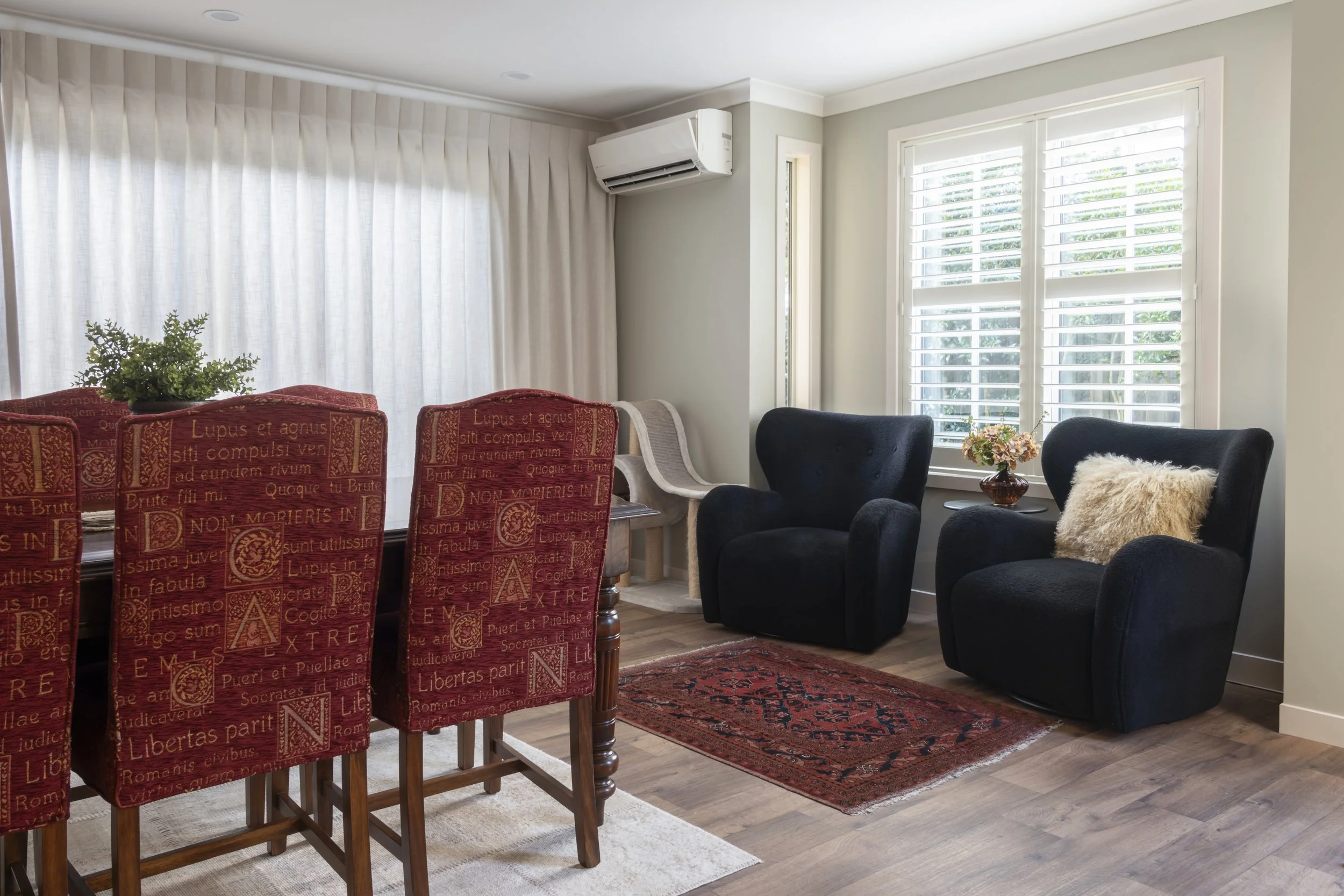 Living room with red dining chairs, black armchairs, large window with white blinds, air conditioning unit, and a small table with a flower vase.