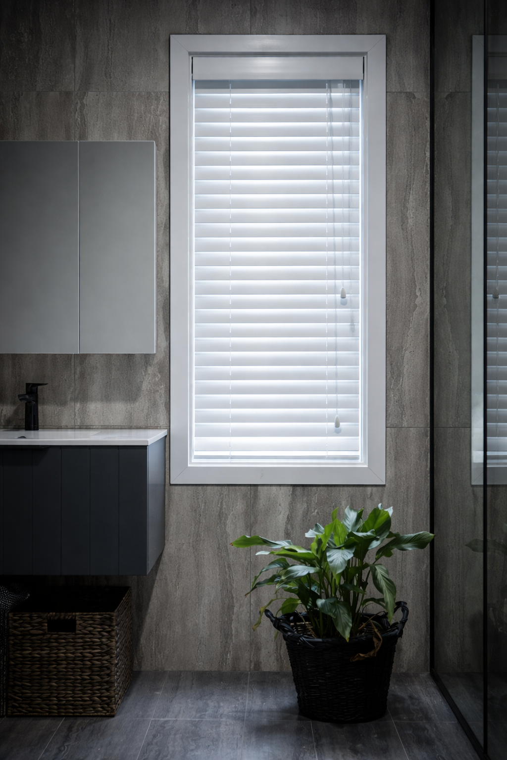 A modern bathroom with a gray cabinet, a potted green plant in a black basket, a window with white blinds, and a textured gray wall.