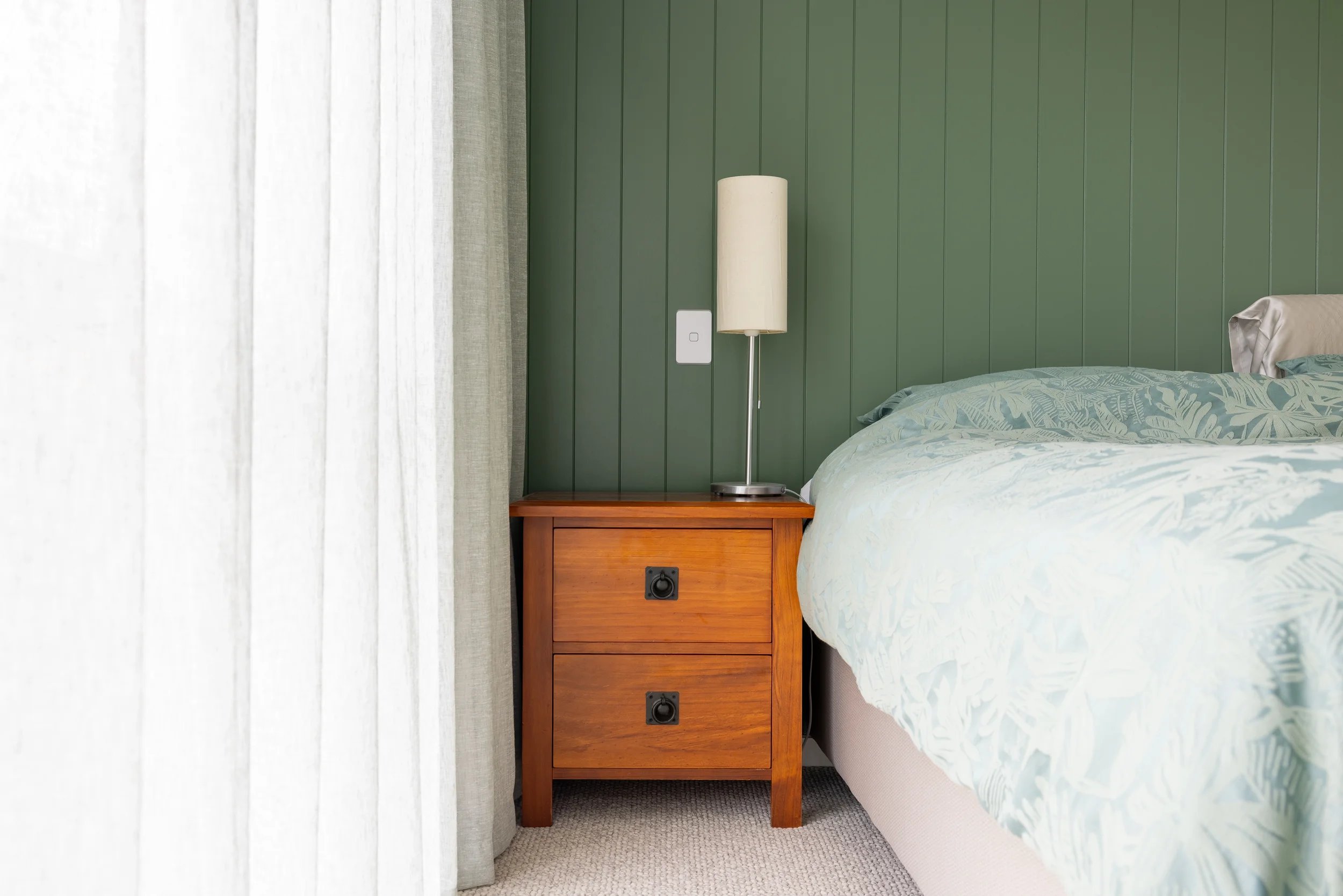 A wooden nightstand with two drawers next to a bed with a light-colored floral bedspread. A tall white lamp sits on the nightstand, and there is a green paneled wall with a white light switch behind it. White curtains cover a window to the left.