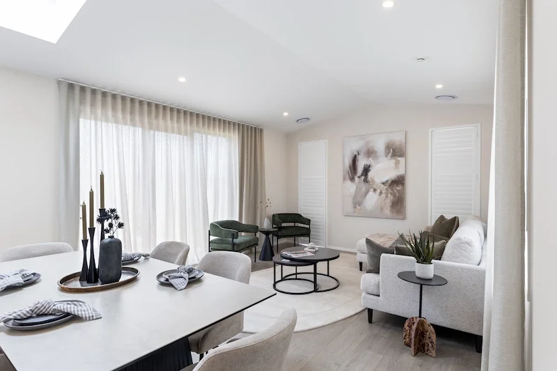 Modern living room and dining area with white walls, light wood flooring, floor-to-ceiling beige curtains, and contemporary furniture including a white sofa, green chairs, and a black round coffee table.