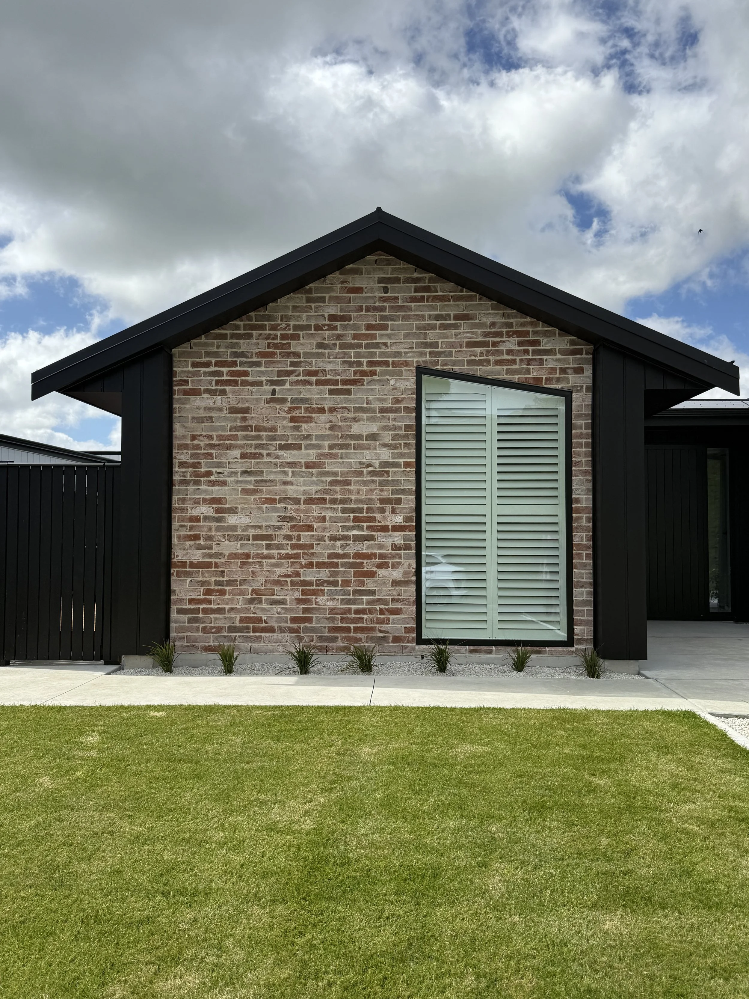 Front view of a modern house with a gabled roof, brick facade, large window with shutters, black siding, concrete pathway, neatly trimmed lawn, and cloudy sky.