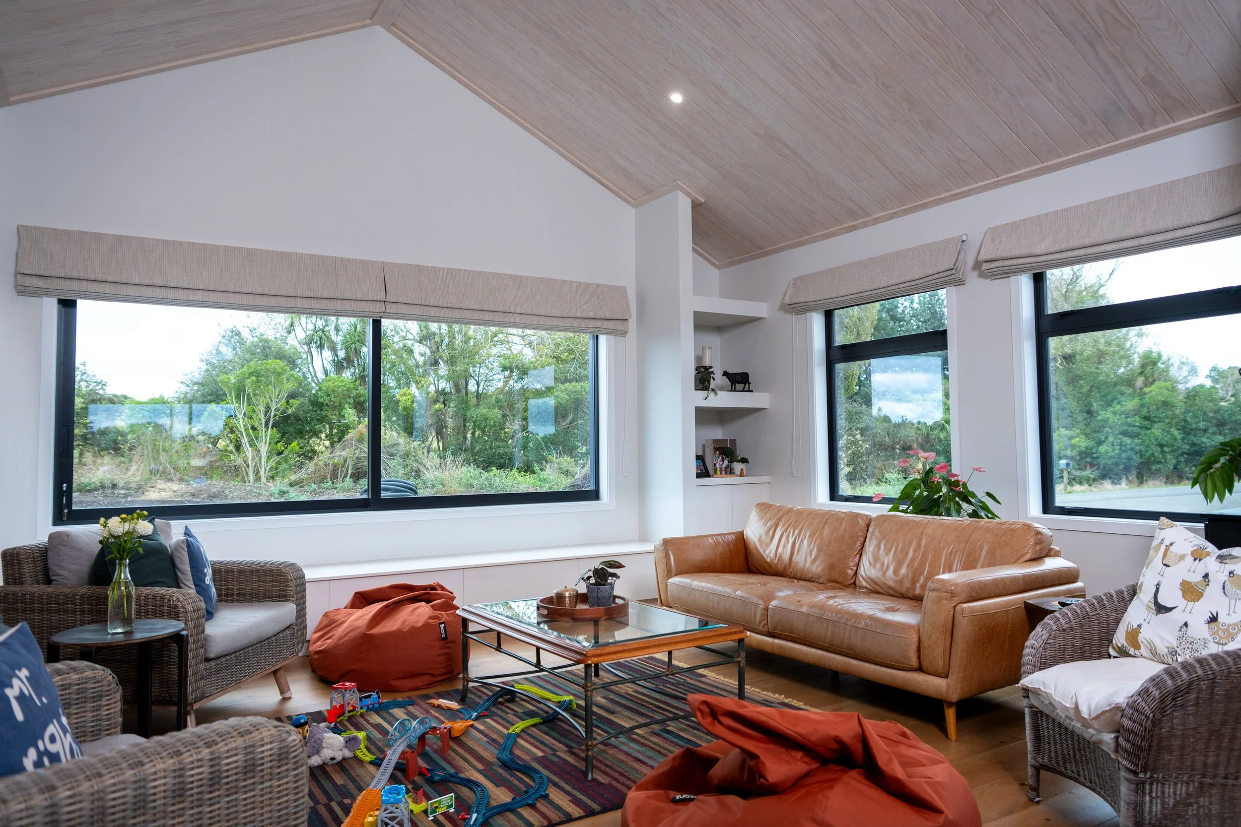 Living room with large windows showing outdoor greenery, tan leather sofa, wicker chairs, a glass coffee table, bean bags, and children's train tracks on a colorful rug.