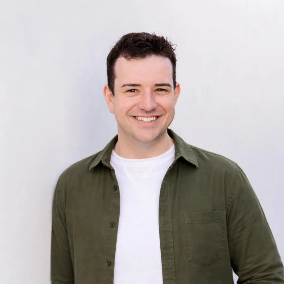 A young man sitting in a modern gray armchair with his hands clasped on his lap, smiling at the camera. He is wearing a green jacket and black pants, set against a backdrop of assorted black and white patterned curtains.
