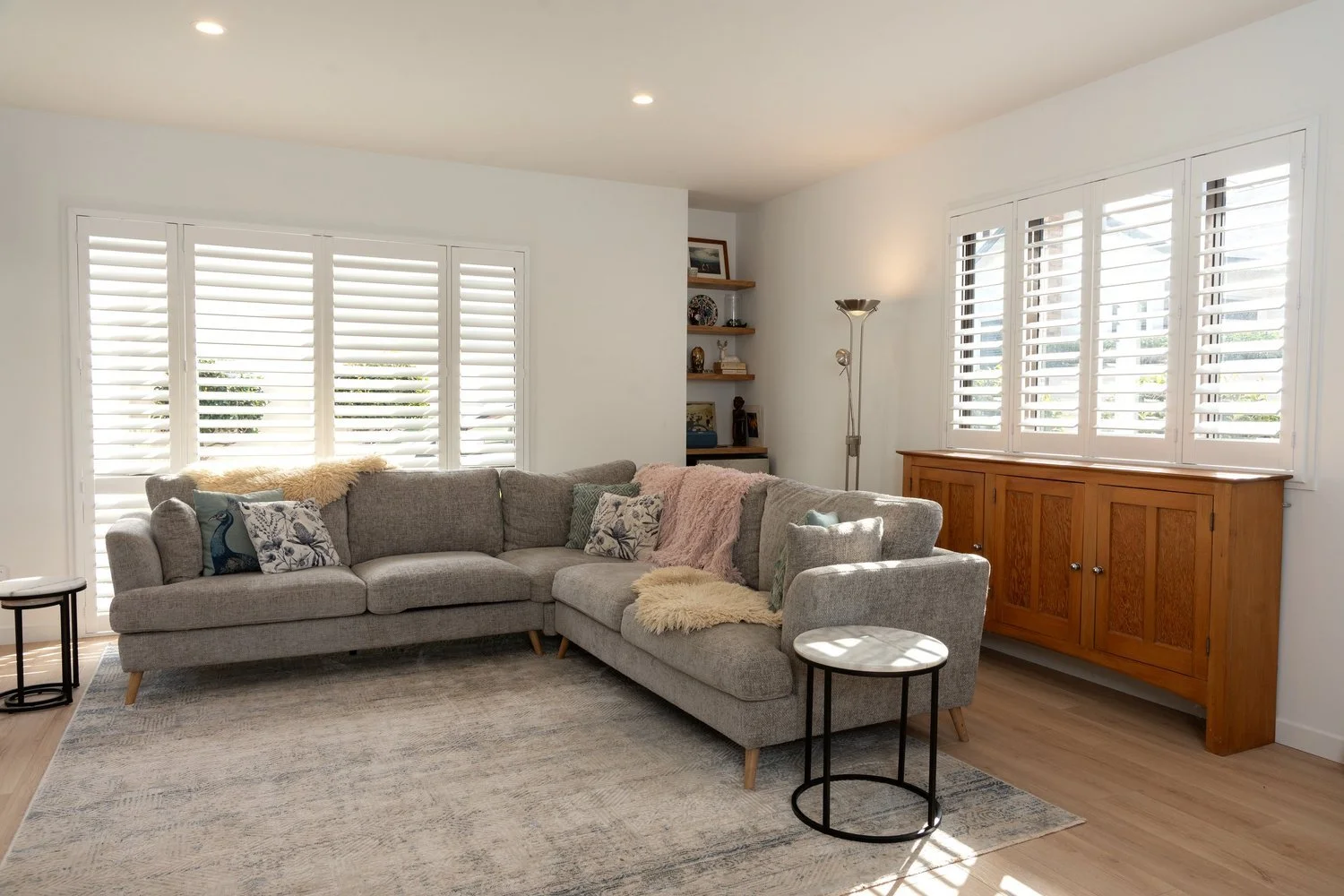 Living room with large gray sectional sofa, decorative pillows, throw blankets, light hardwood floor, window shutters, built-in wooden cabinet, and small side tables.