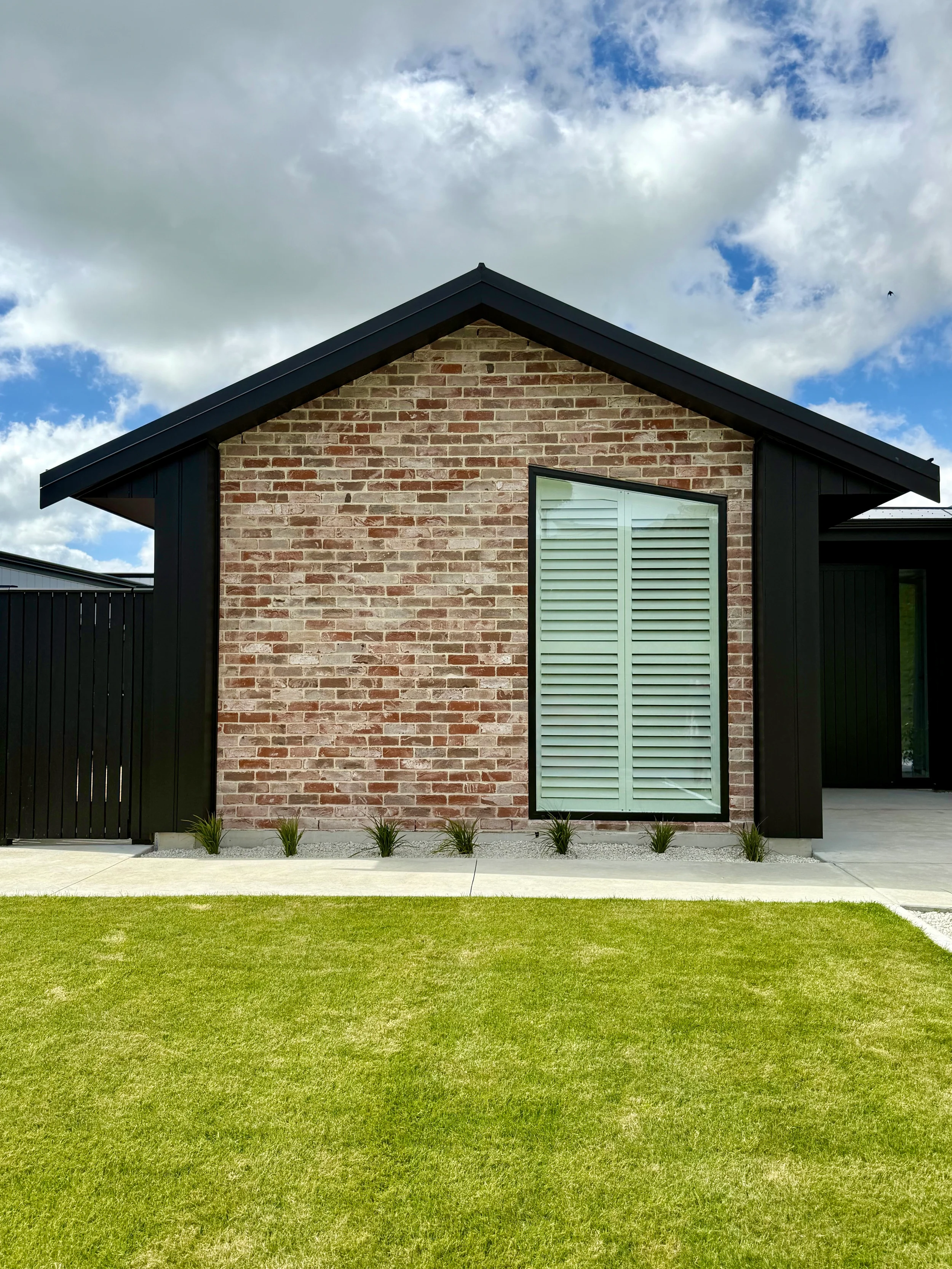 Modern house with brick and black siding, large window with shutter, small plants at the base, grass, and a partly cloudy sky.