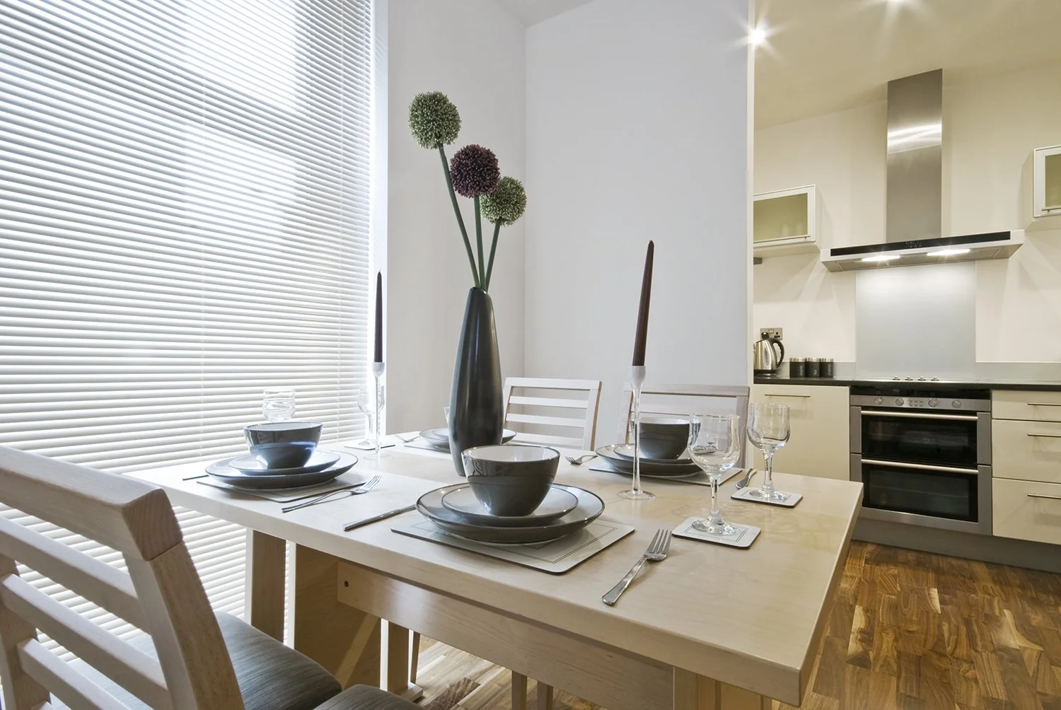 A modern dining room with a wooden table set for four, featuring gray bowls, plates, and glasses, with a dark vase holding large decorative flowers, adjacent to a white kitchen with stainless steel appliances and hardwood floors.