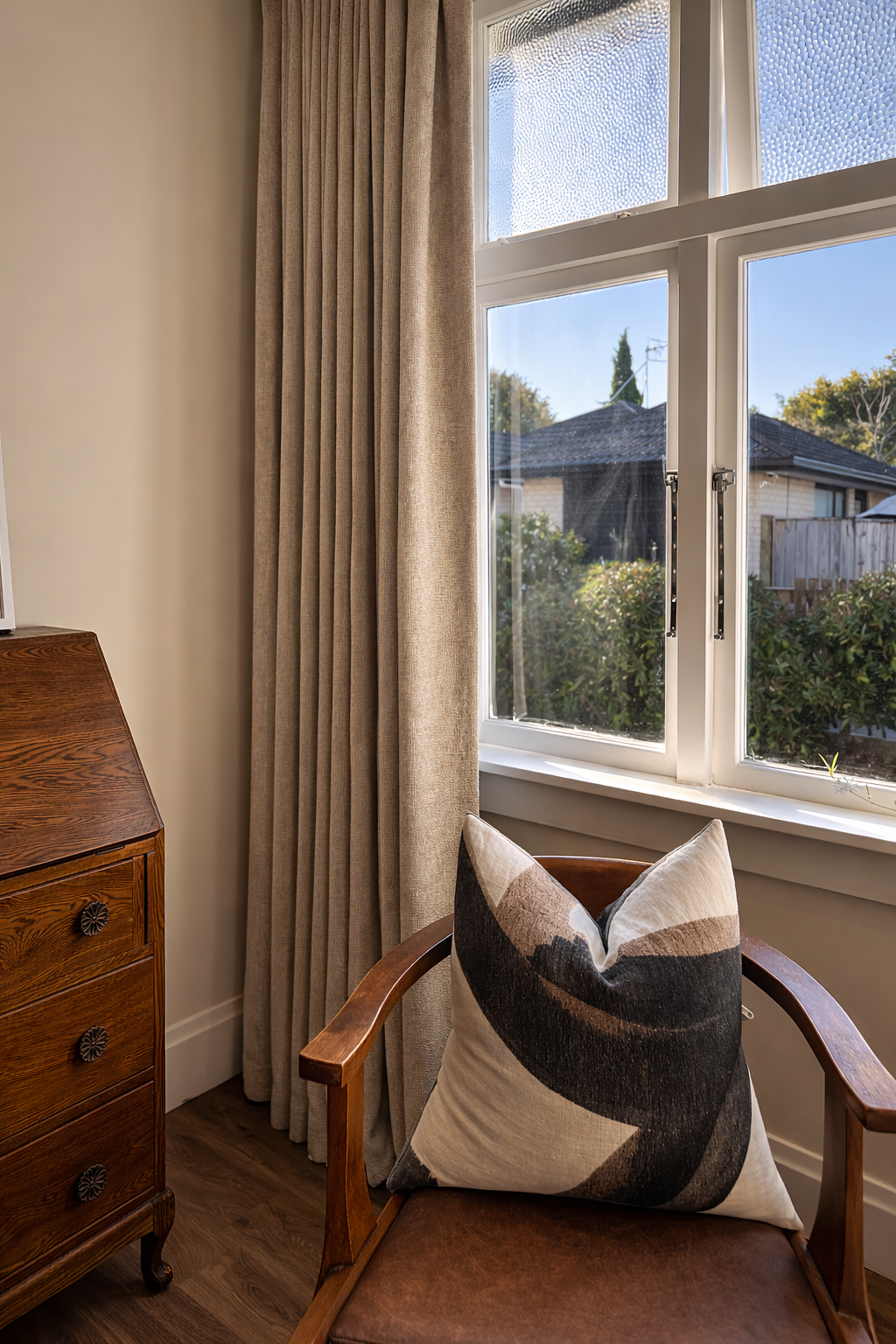A cozy corner of a room with beige curtains, a wooden dresser with round black knobs, a wooden chair with a brown leather seat, and a decorative pillow with black, brown, beige, and white abstract patterns. Outside the window, there are bushes and a 