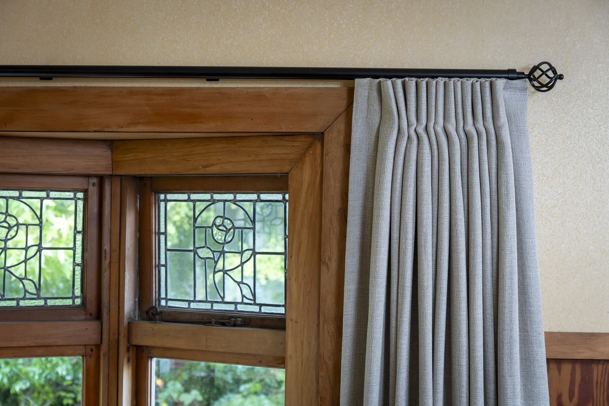 Close-up of a window with wooden framing, stained glass details, and a beige curtain hanging from a black curtain rod.