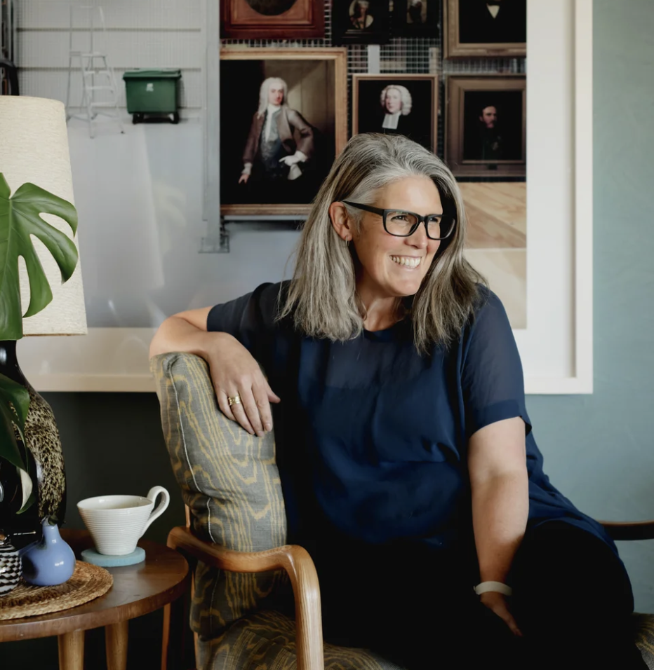 Smiling woman with gray hair and glasses sitting in a chair in a room with framed portrait paintings on the wall behind her.