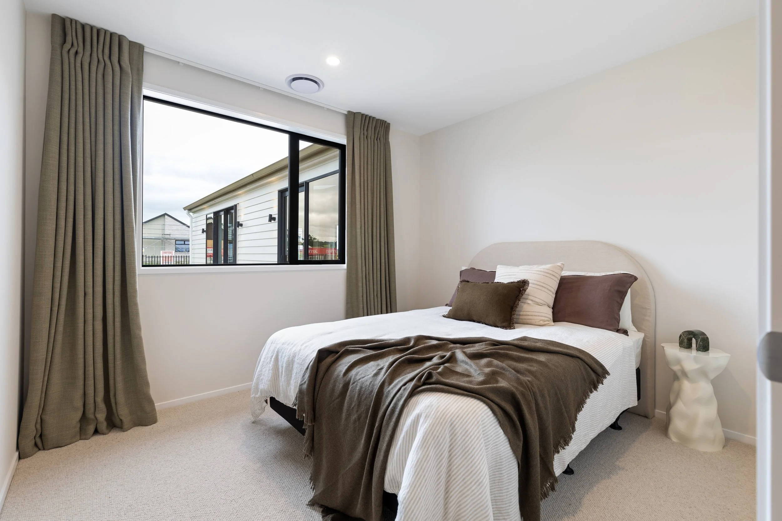 A simple bedroom with a bed, pillows, and a nightstand, featuring a window with curtains showing neighboring houses.