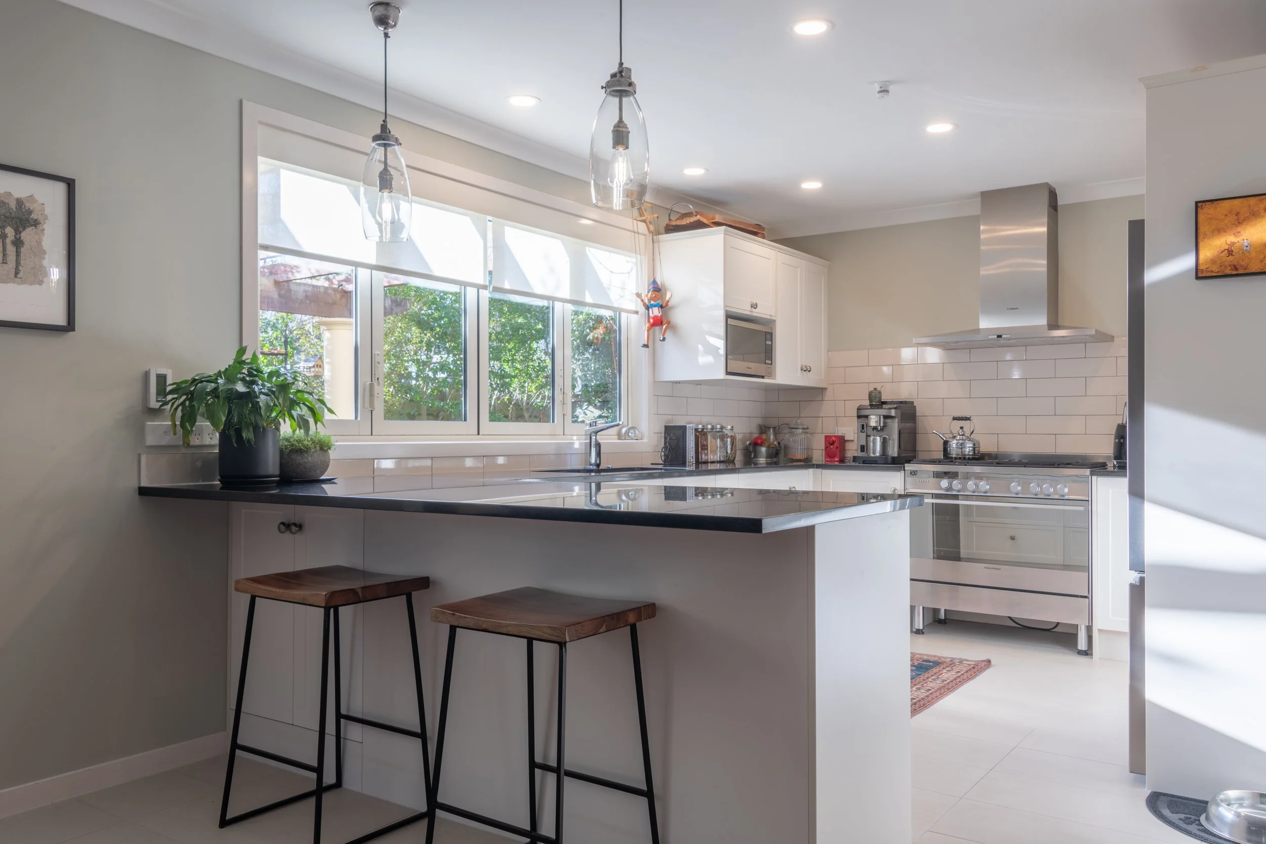 Modern kitchen with white cabinets, black countertop, pendant lighting, window, plants, and stainless steel appliances.