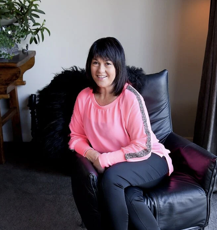 Woman wearing sunglasses and a red and white checkered shirt, smiling outdoors in a garden with trimmed bushes and a building in the background.