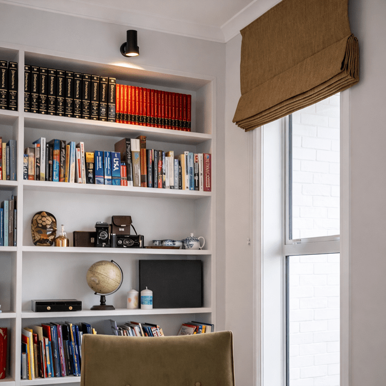 A white bookshelf filled with books, decorative items, and cameras next to a window with a brown fabric blind in a room with white walls.