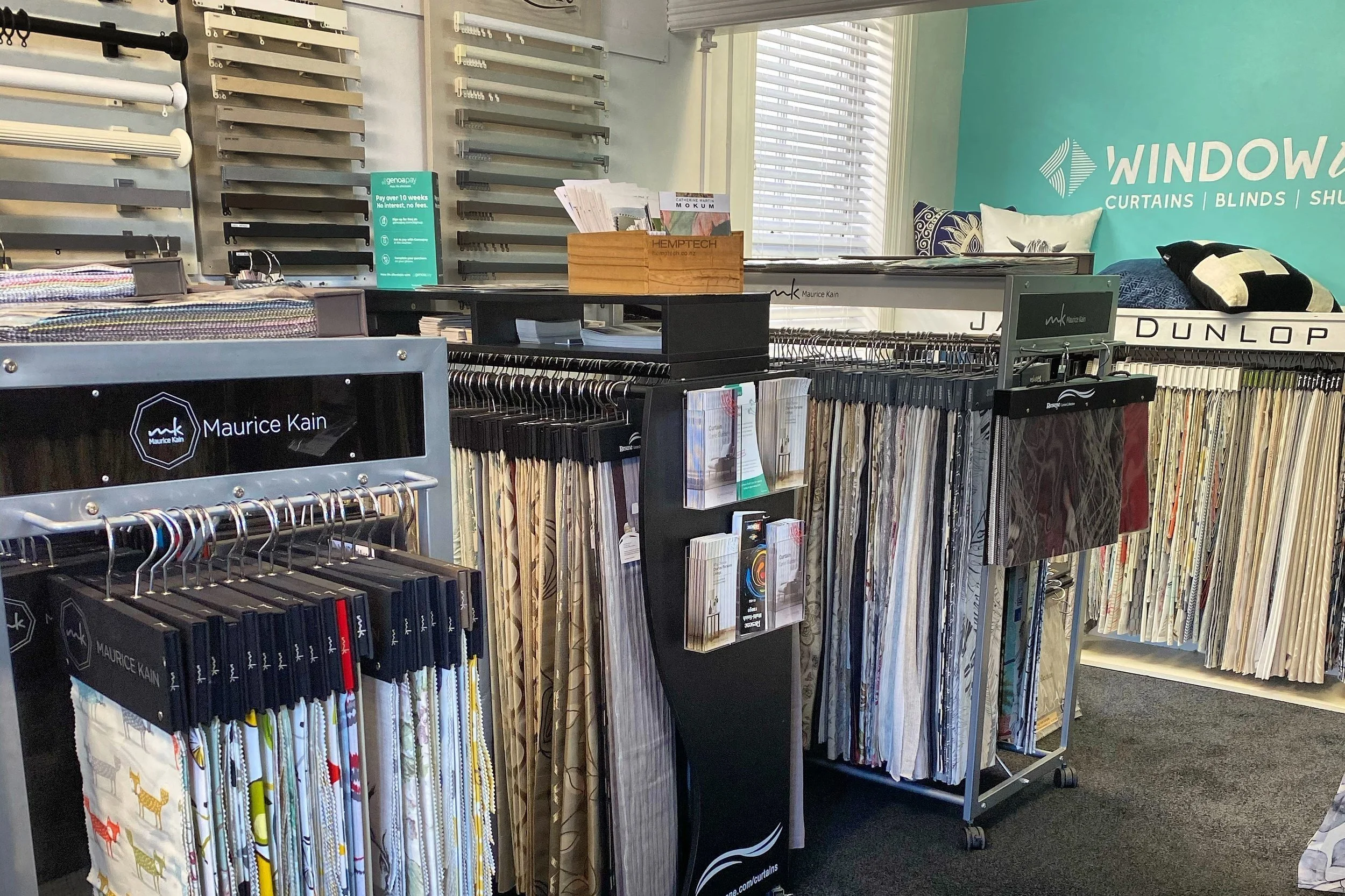 Indoor fabric store display featuring curtains and blinds, with fabric samples on racks, signage for Maurice Kain and Dunlop, and a teal wall with window-themed branding.