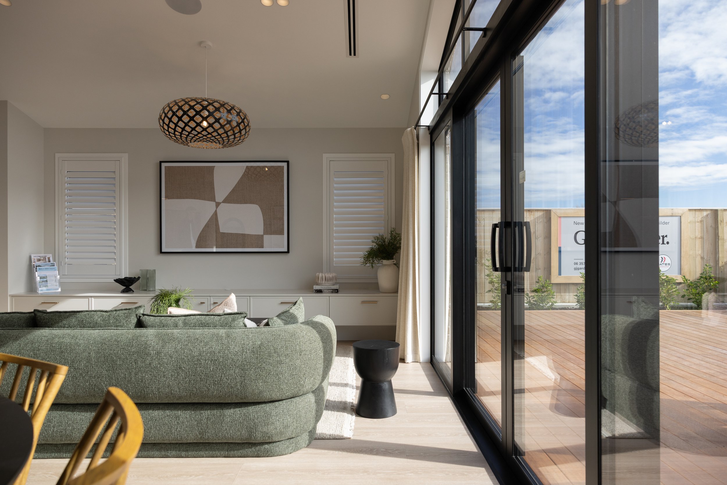 Interior view of a modern living room featuring a green sofa, a large framed abstract art on the wall, a potted plant, and a black side table. Floor-to-ceiling black-framed glass sliding doors open to a wooden deck outside, with a partly cloudy sky v
