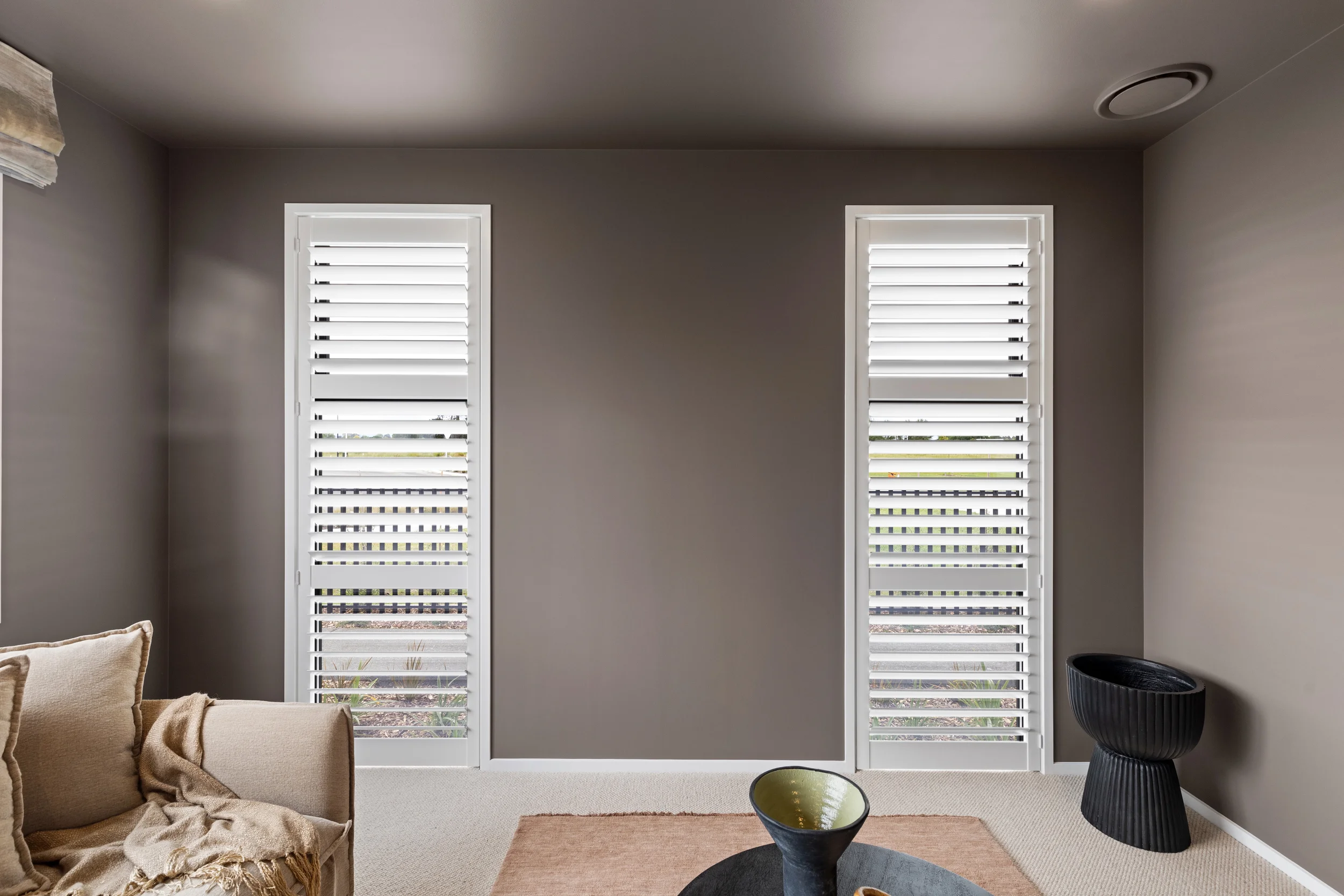 A modern living room with two tall windows with white plantation shutters, a beige sofa with cushions, a black textured side table, a small round black coffee table with a decorative bowl, and a light-colored carpet. Gray walls and ceiling, with minimal decor.