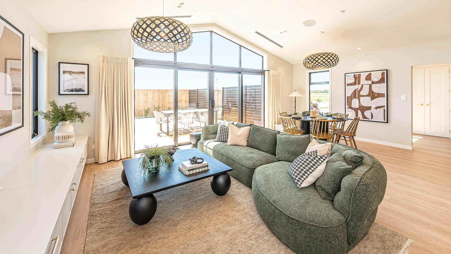 Living room with large glass sliding doors leading to an outdoor patio, green sectional sofa with throw pillows, black coffee table, white console with decorative items, framed artwork on walls, wooden dining table with chairs, and modern light fixtures.