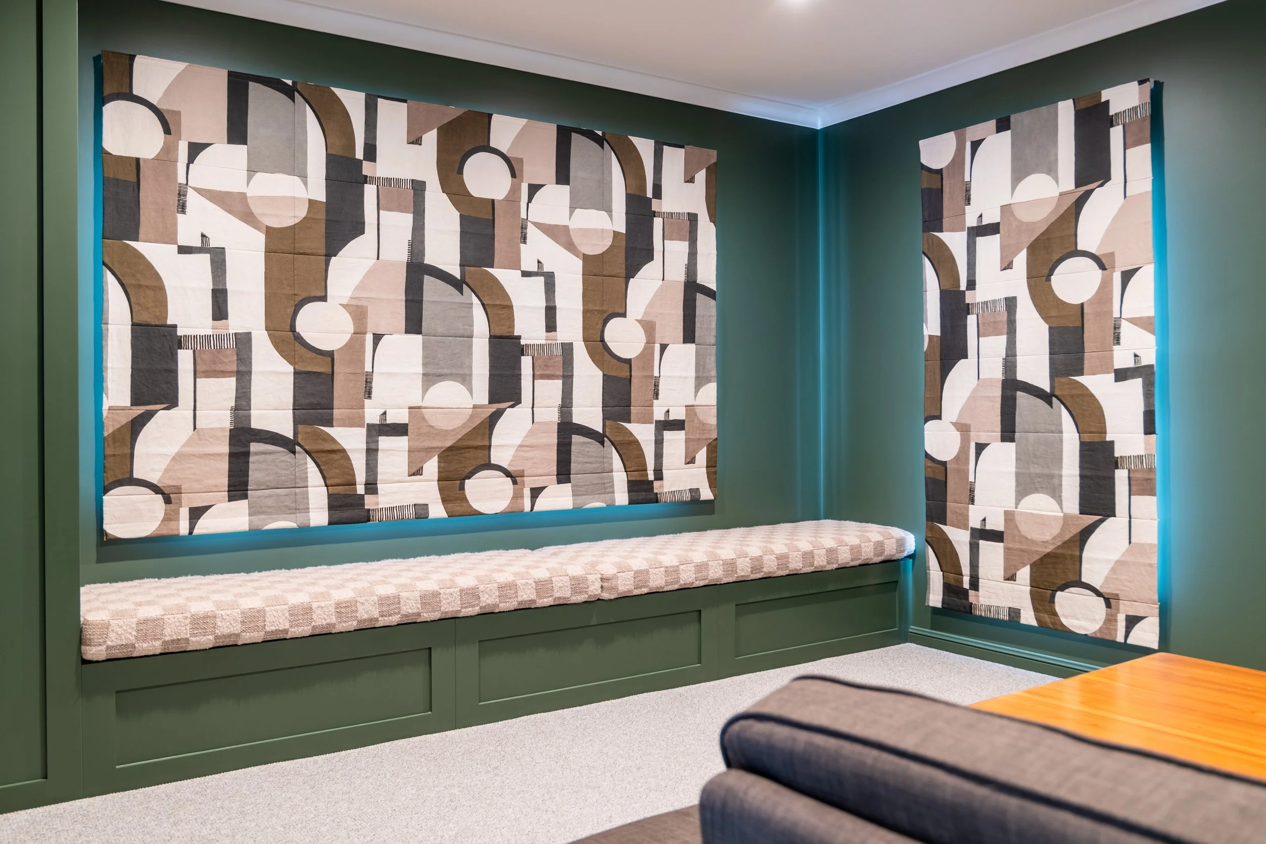 Living room corner with green walls, geometric patterned curtains, a built-in bench with a beige cushion, and a partial view of an orange table and gray sofa.