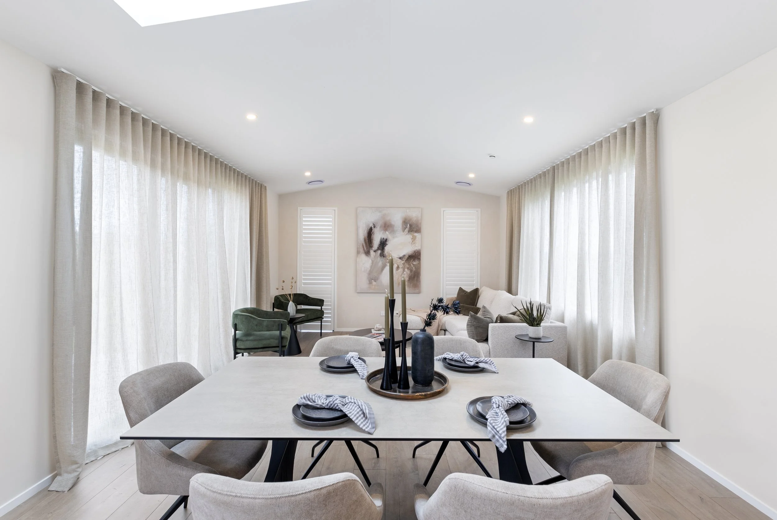 Modern dining and living room with white walls, beige curtains, a rectangular dining table with six beige chairs, and a cozy sitting area with a white sofa, green chairs, and abstract wall art.