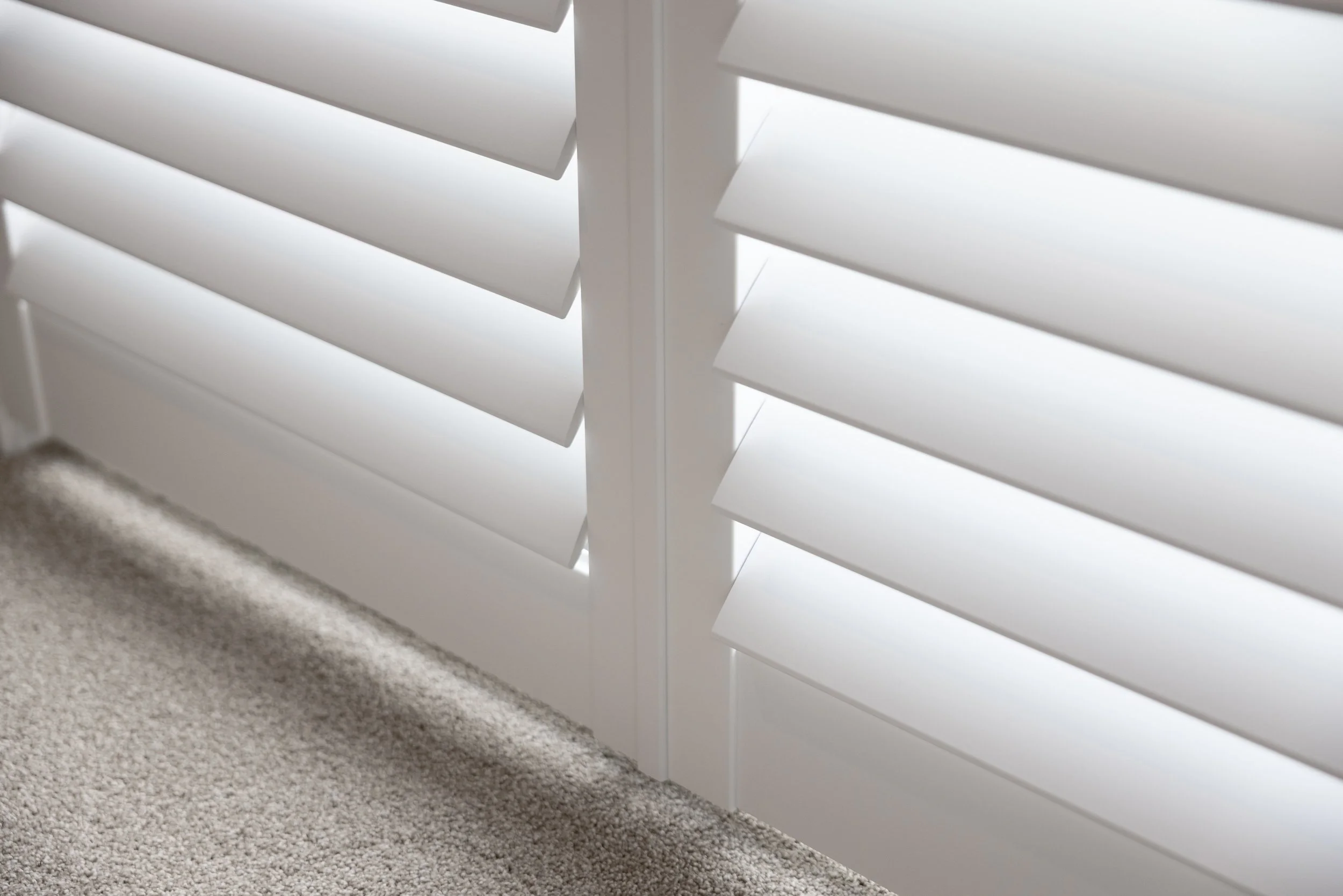 Close-up of white window shutters with partially closed slats, sunlight shining through, next to beige carpeted flooring.