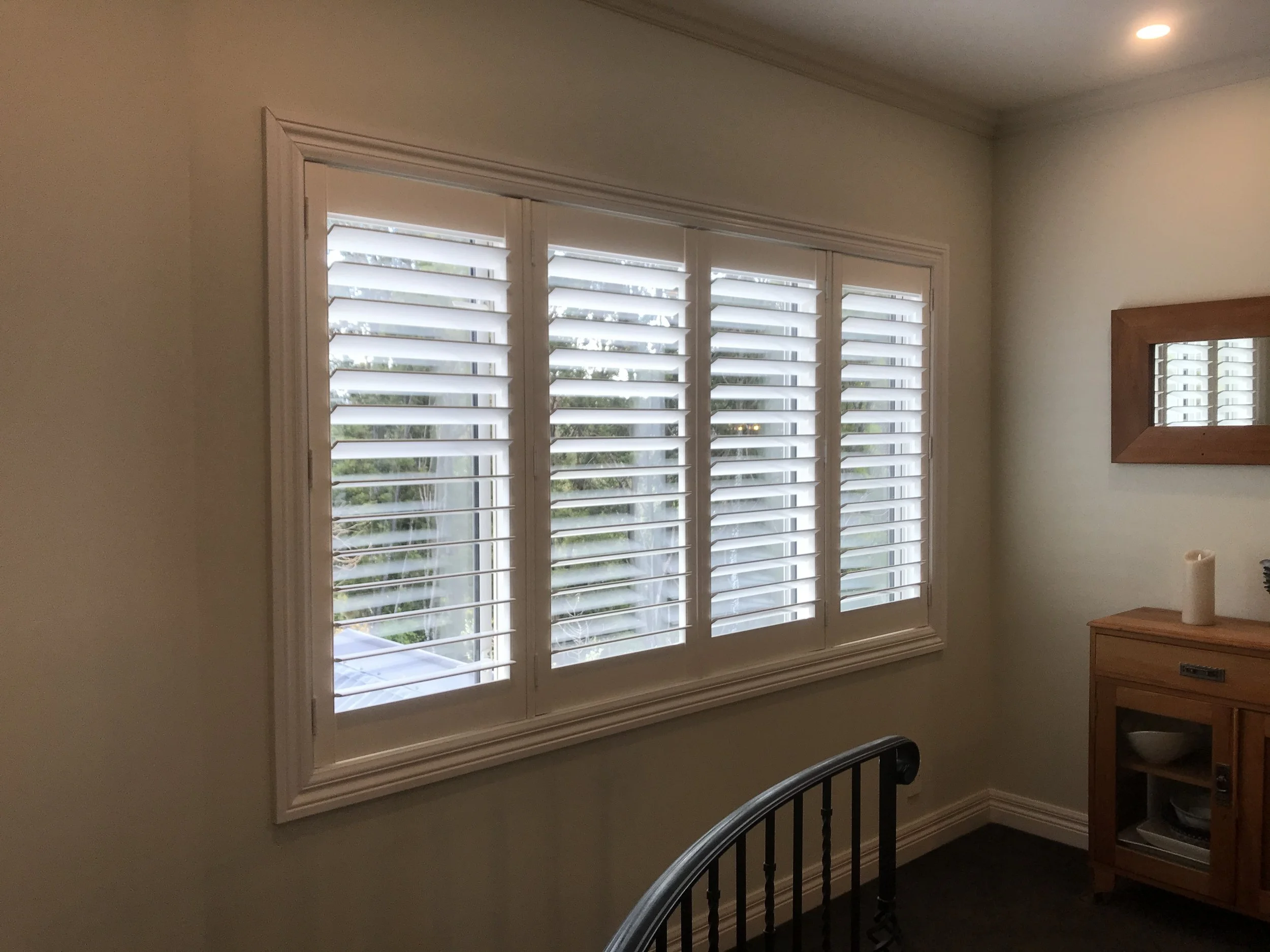 Room with three-panel window with white plantation shutters and a wooden sideboard with a mirror above it.