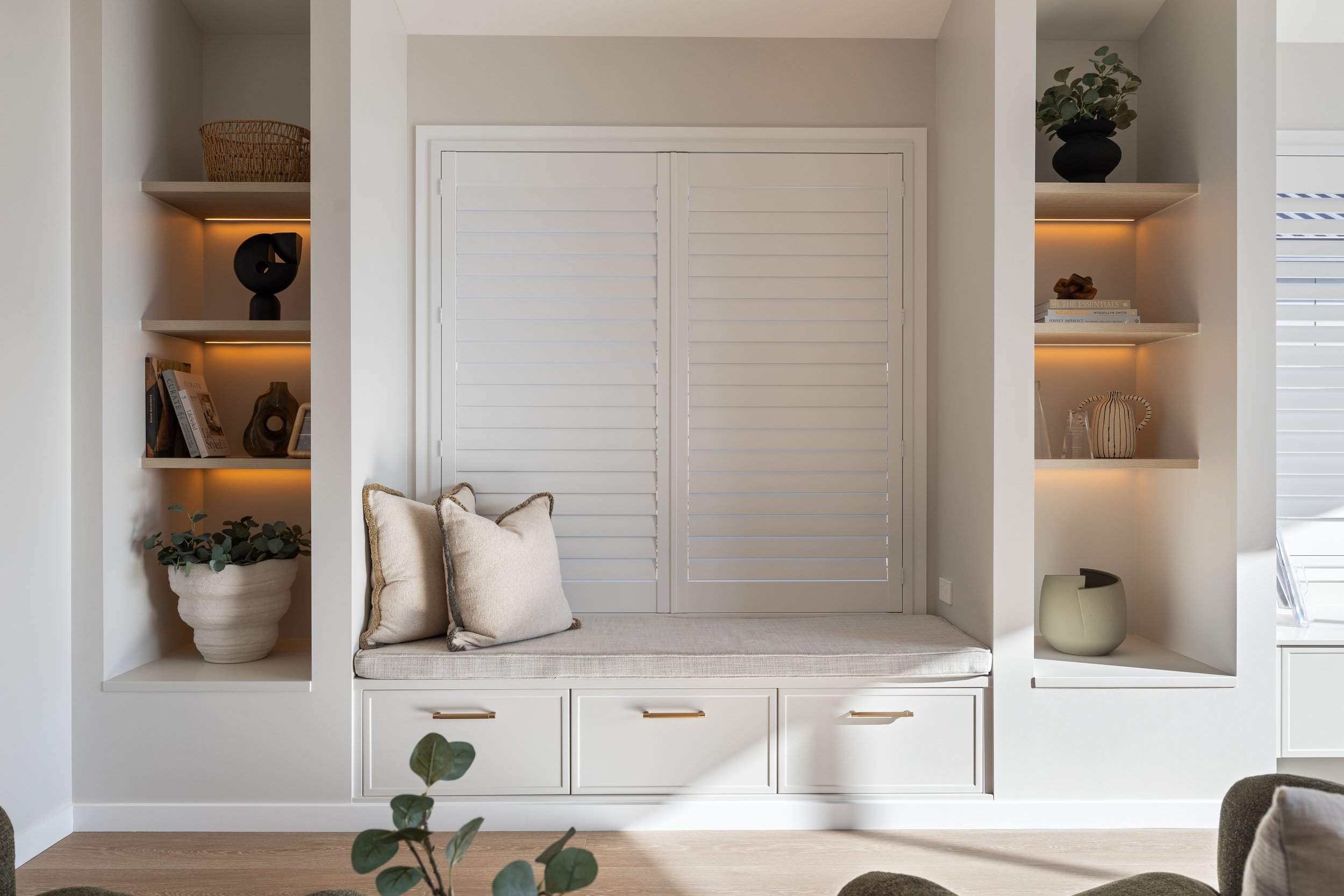 Built-in white shelving unit with books, decorative objects, and plants, featuring a small seating area with cushions.
