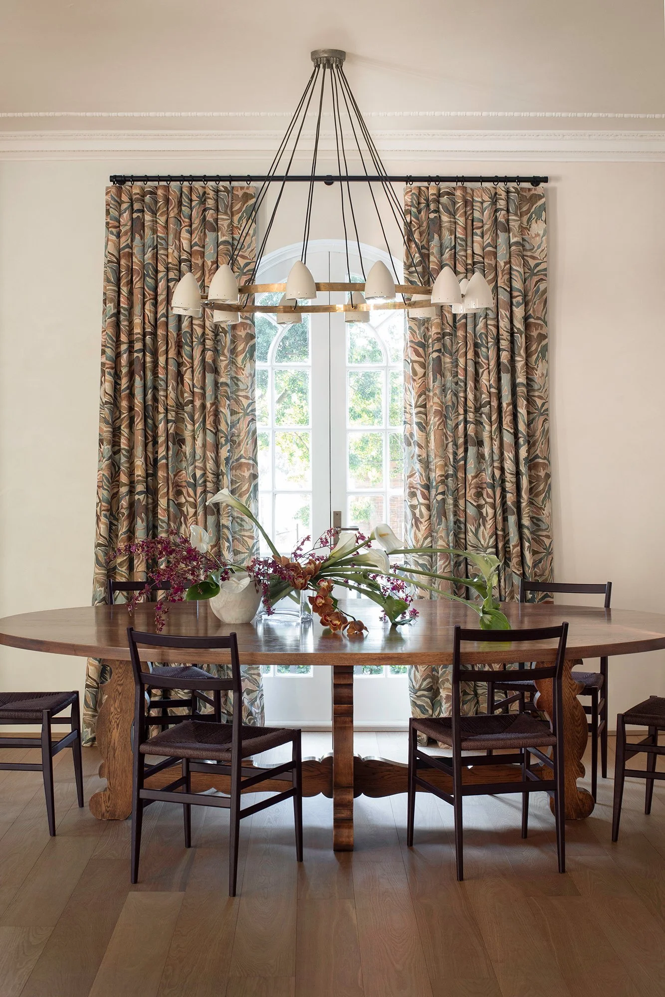 A dining room with a wooden table, decorated with flower arrangements in a vase, surrounded by black chairs. Behind the table, there are large windows with floral patterned curtains, and a modern chandelier hanging from the ceiling.