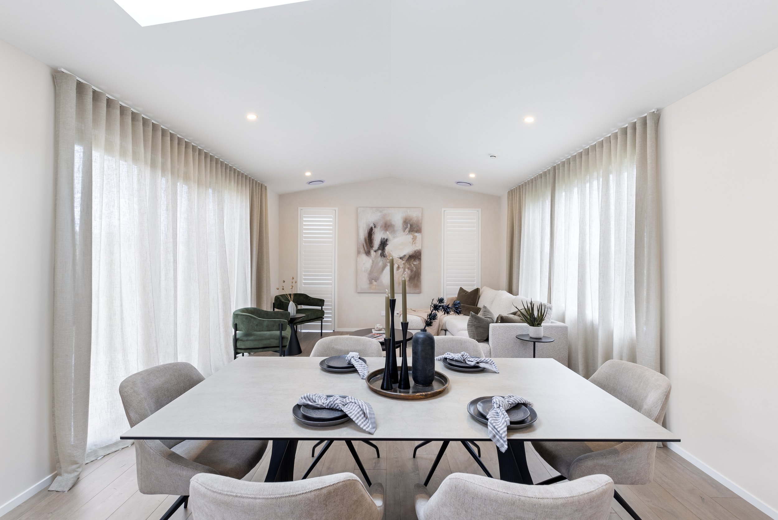 Modern living and dining room with beige chairs around a dining table, white sofa with pillows, abstract wall art, and large windows with sheer curtains.
