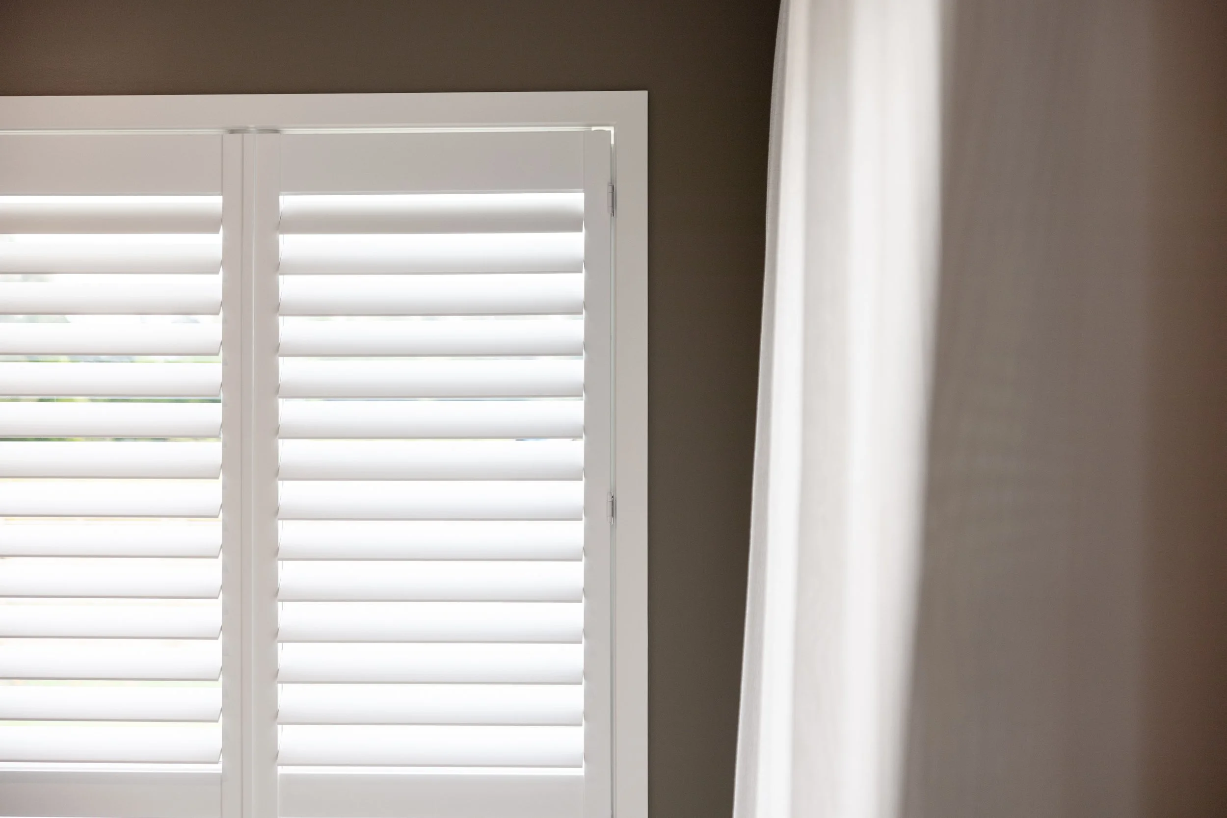Part of a window with white plantation shutters and a beige curtain on the right side.