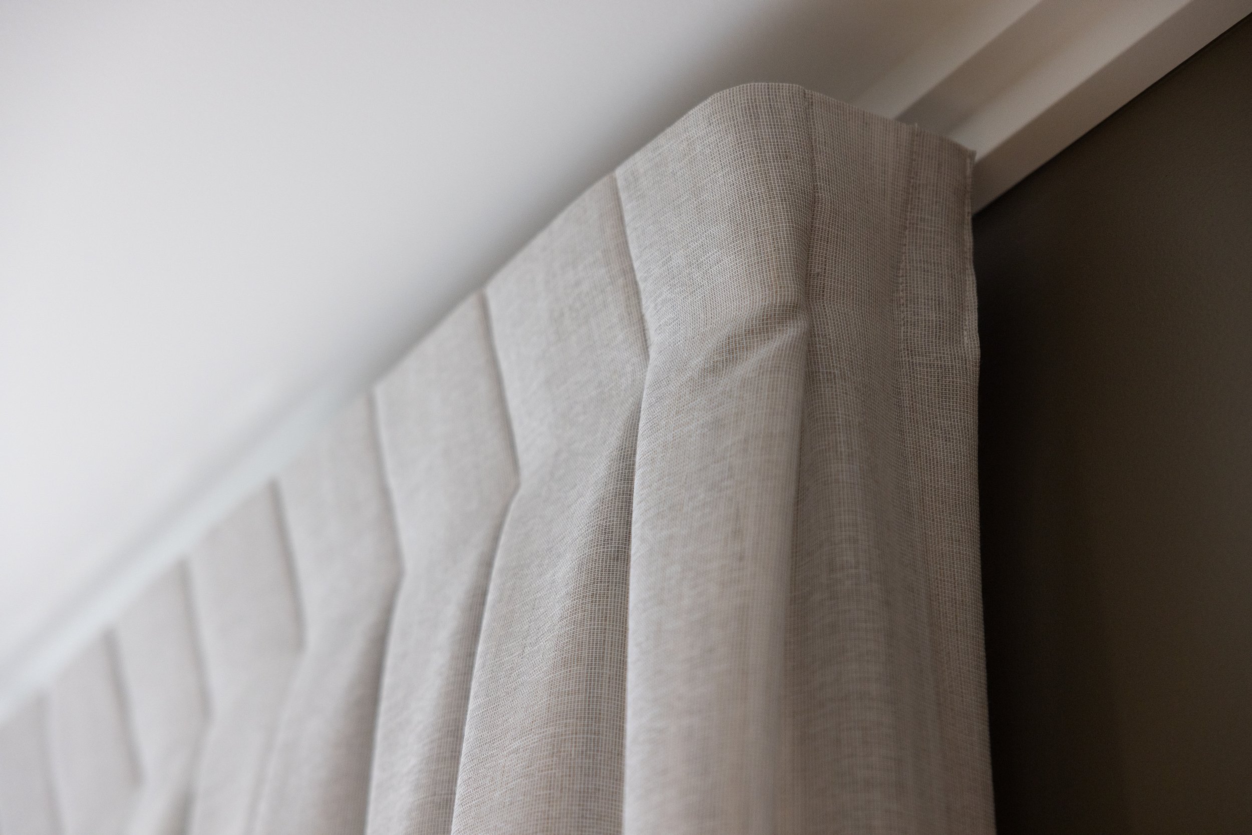 Close-up of a beige upholstered headboard with a piece of sheer curtain hanging beside it.
