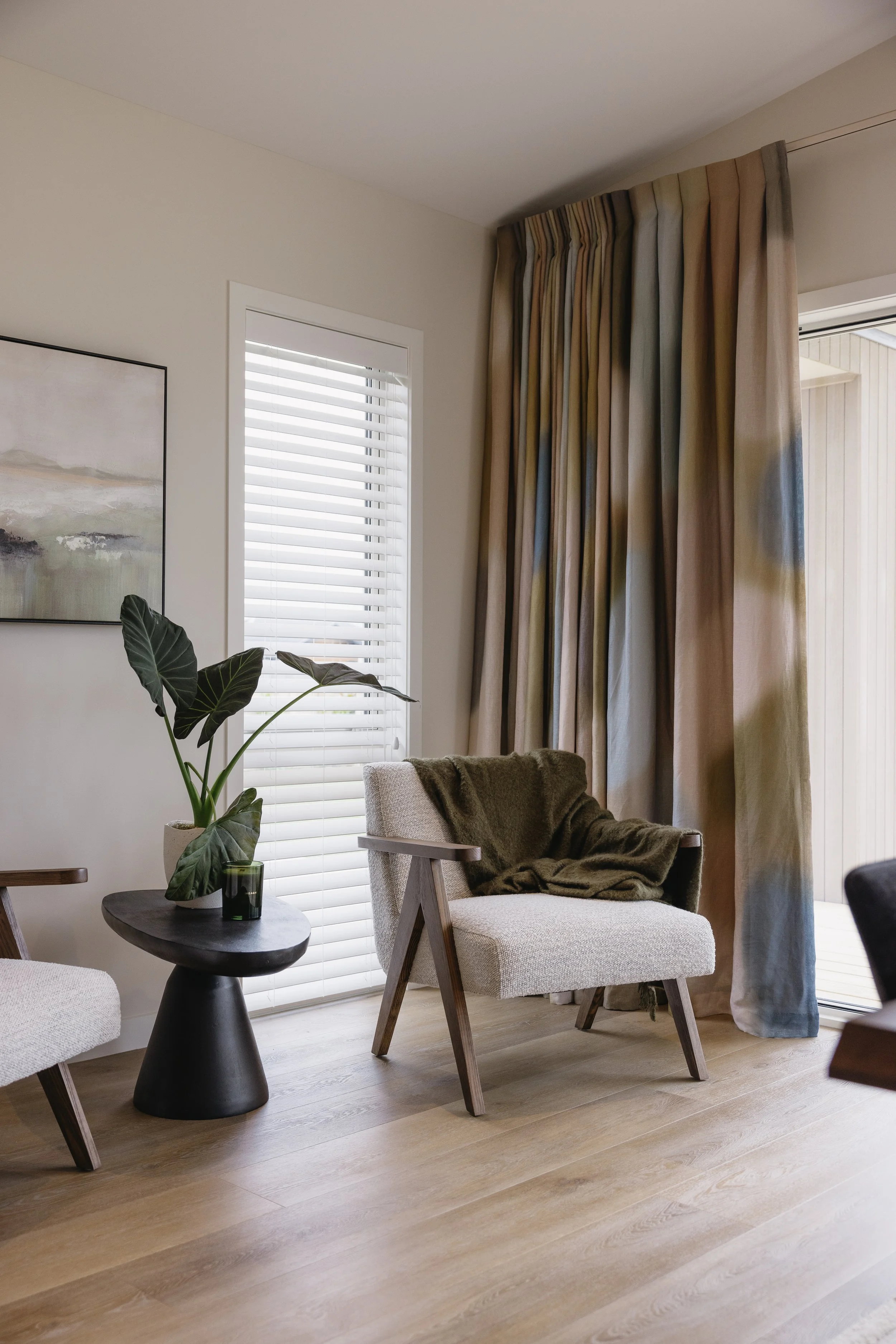 A cozy living room corner featuring a beige upholstered armchair with a brown blanket draped over it, a black side table with a green plant in a white pot, and a small dark-colored candle holder. There is a window with white horizontal blinds and a large window with beige and multicolored curtains. Part of a painting with soft colors is visible on the left side of the image.
