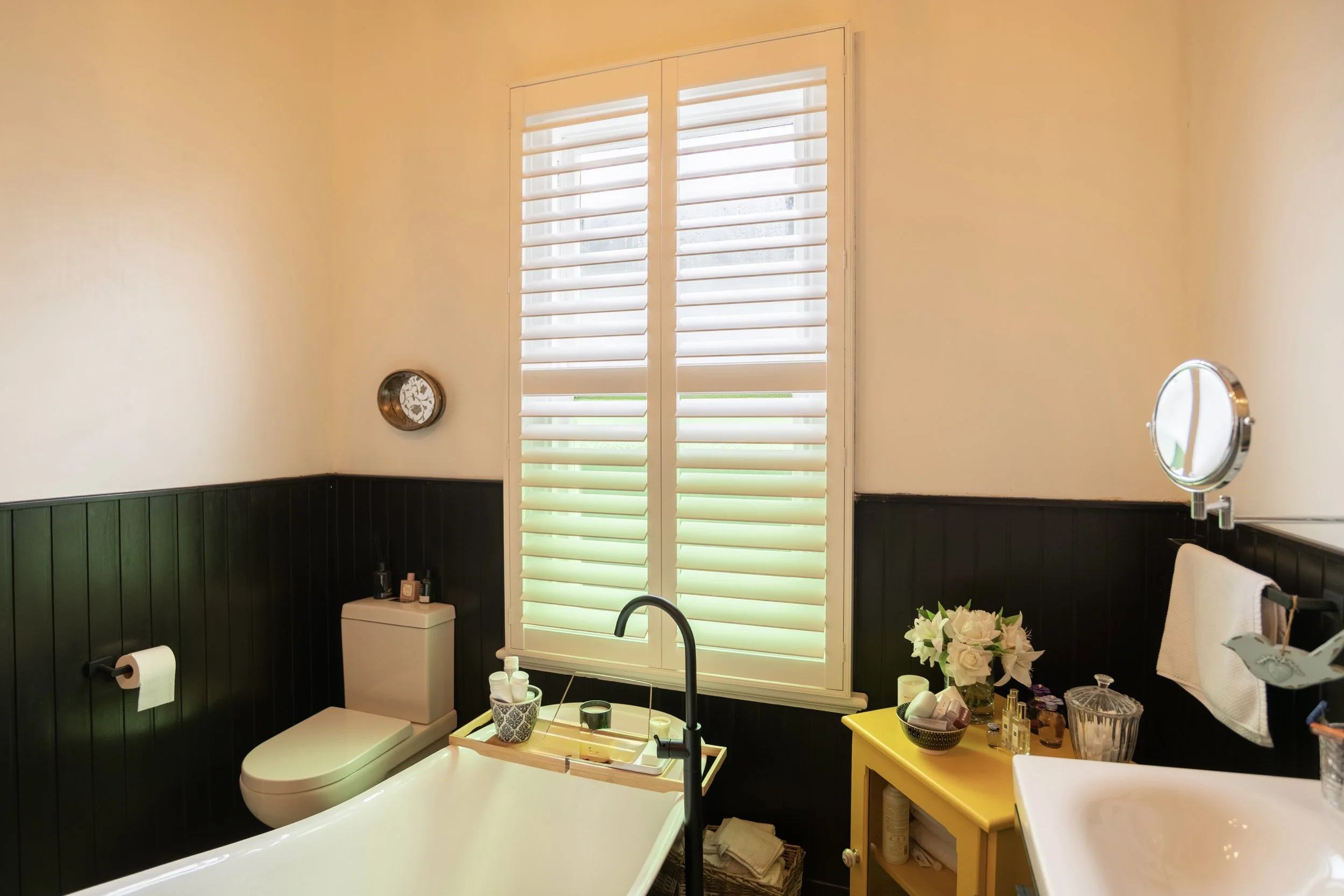 A bathroom with white walls and black wood paneling. It has a white bathtub with a black faucet, a toilet with a small tray on top, a yellow cabinet with decorative items, and a white sink with a round mirror. There is a large window with white shutt