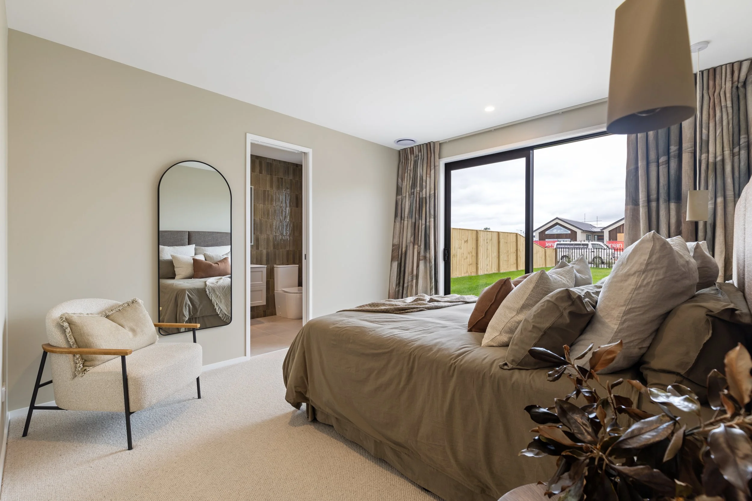 A modern bedroom with a large bed featuring multiple pillows, a beige armchair, a full-length mirror, and a sliding glass door leading to a backyard with a wooden fence and neighboring houses.