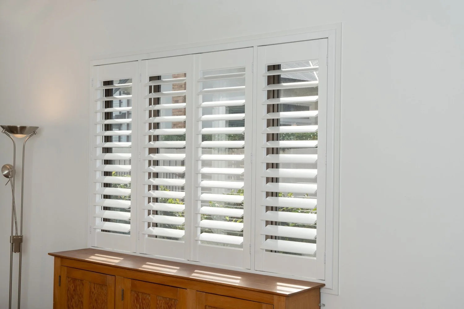 White window with plantation shutters in a bright room, a wooden cabinet below, and a tall floor lamp on the left.