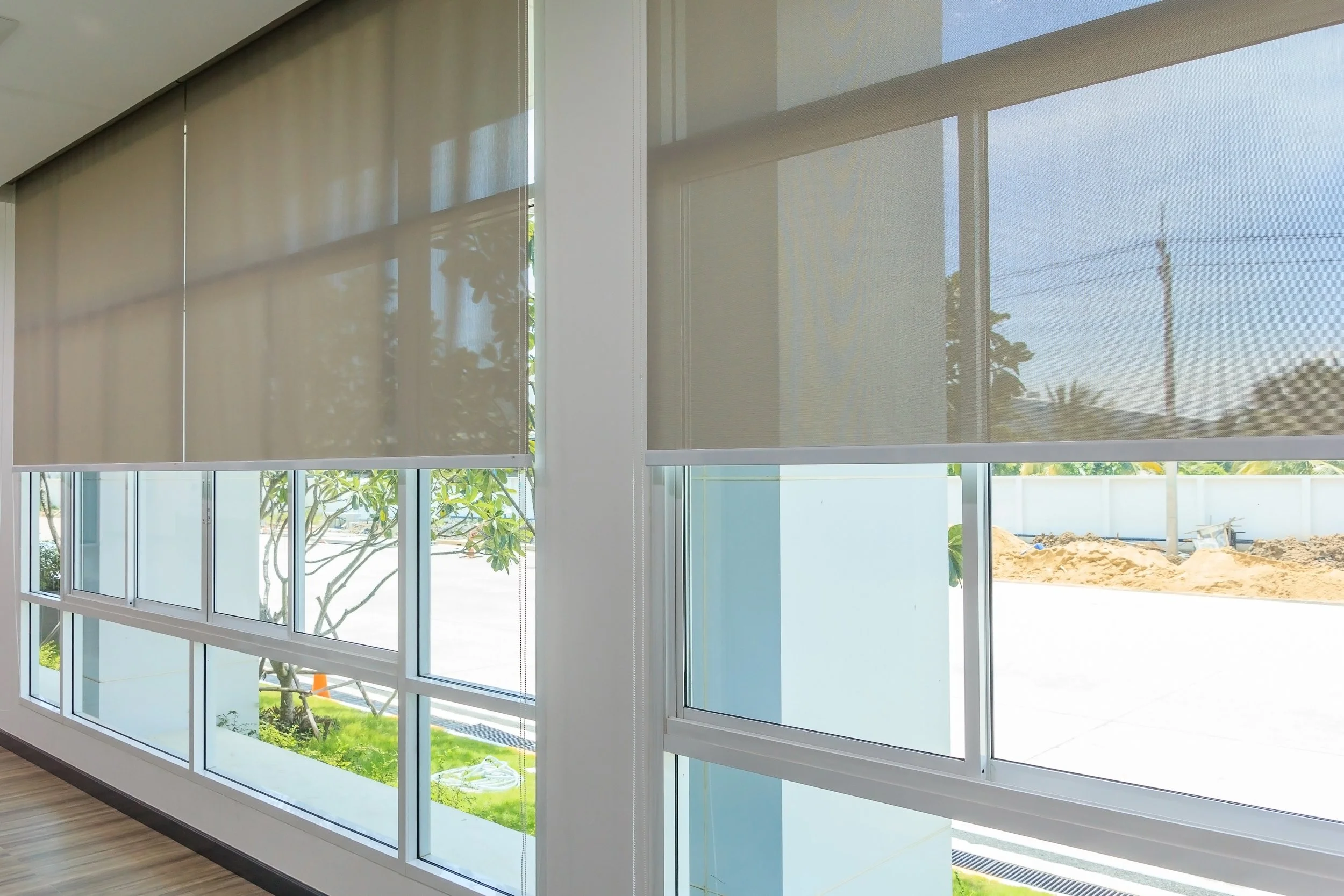 Interior view of large windows with partly closed blinds, showing an outdoor scene with trees, blue sky, and construction dirt.